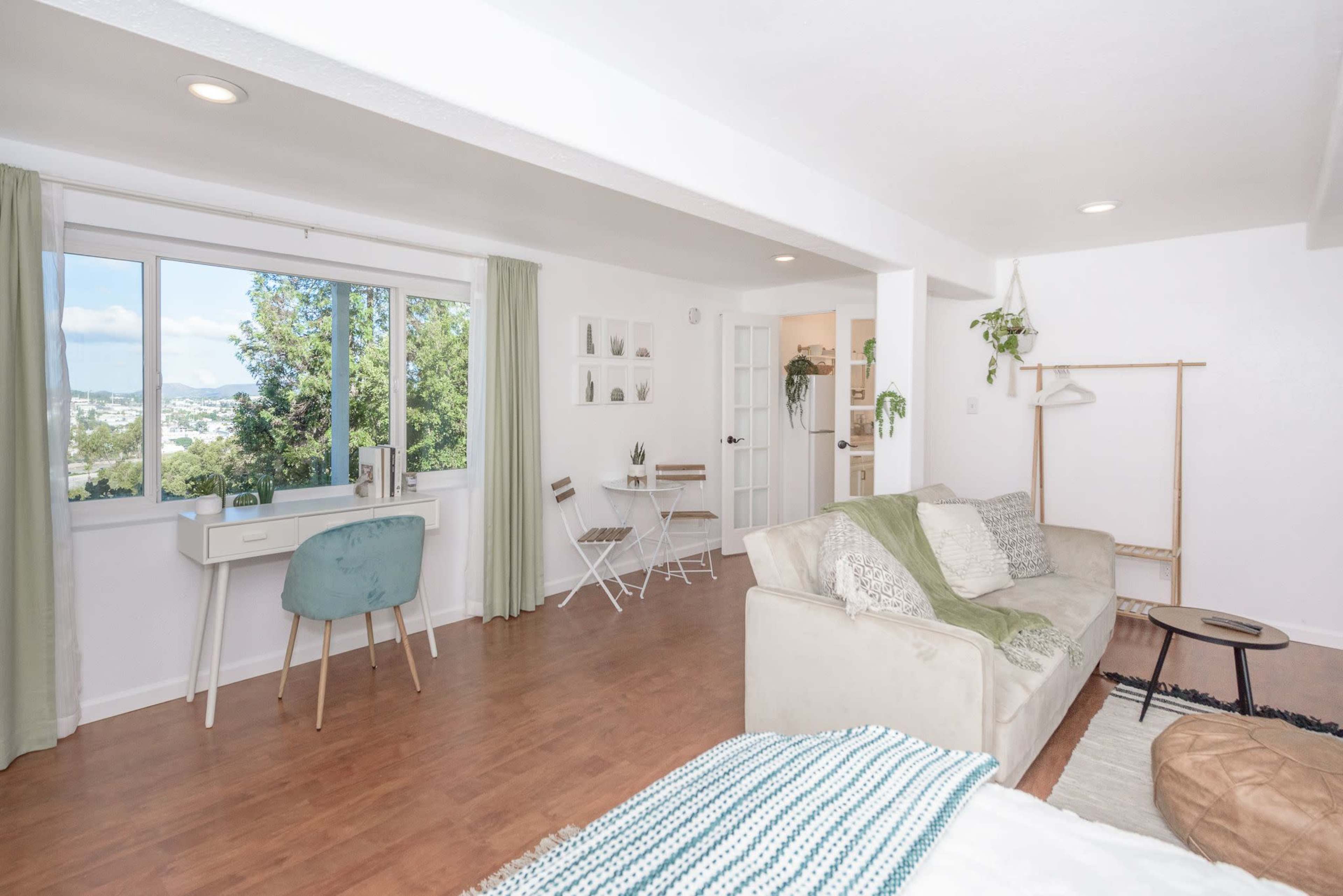 A bright, airy living space features a sofa, a desk by a window, a small dining area with two chairs, and indoor plants.