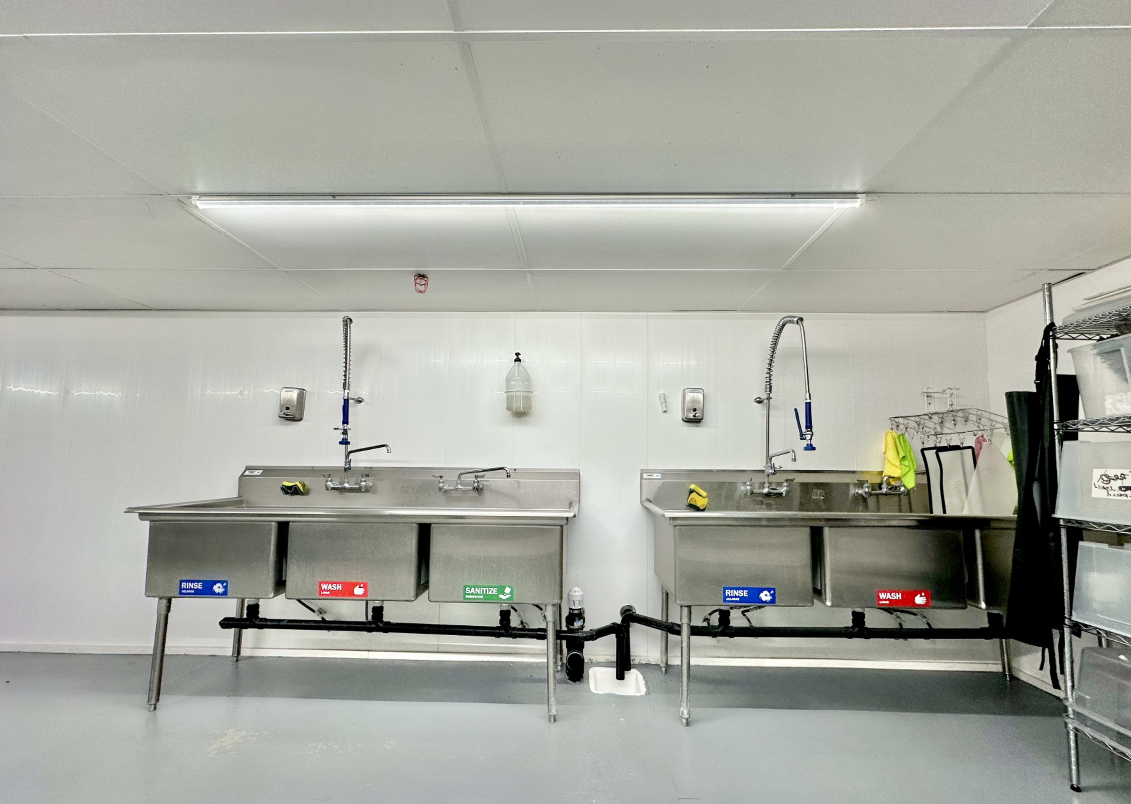 The image shows a clean, well-lit room with two stainless steel sink stations equipped with faucets and color-coded signage underneath each sink.