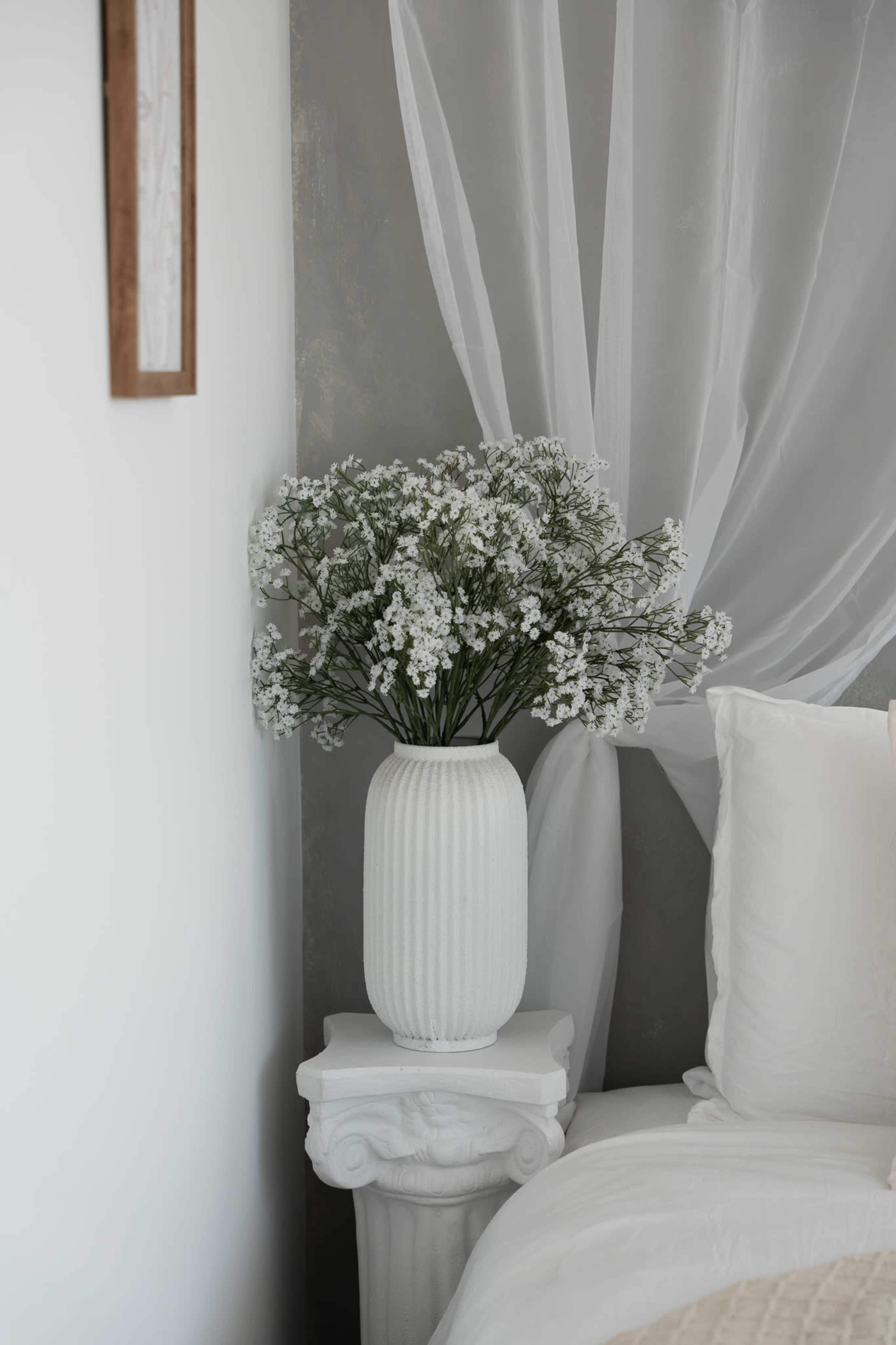 A white vase filled with small white flowers sits on a decorative pedestal next to a bed draped with white linens.