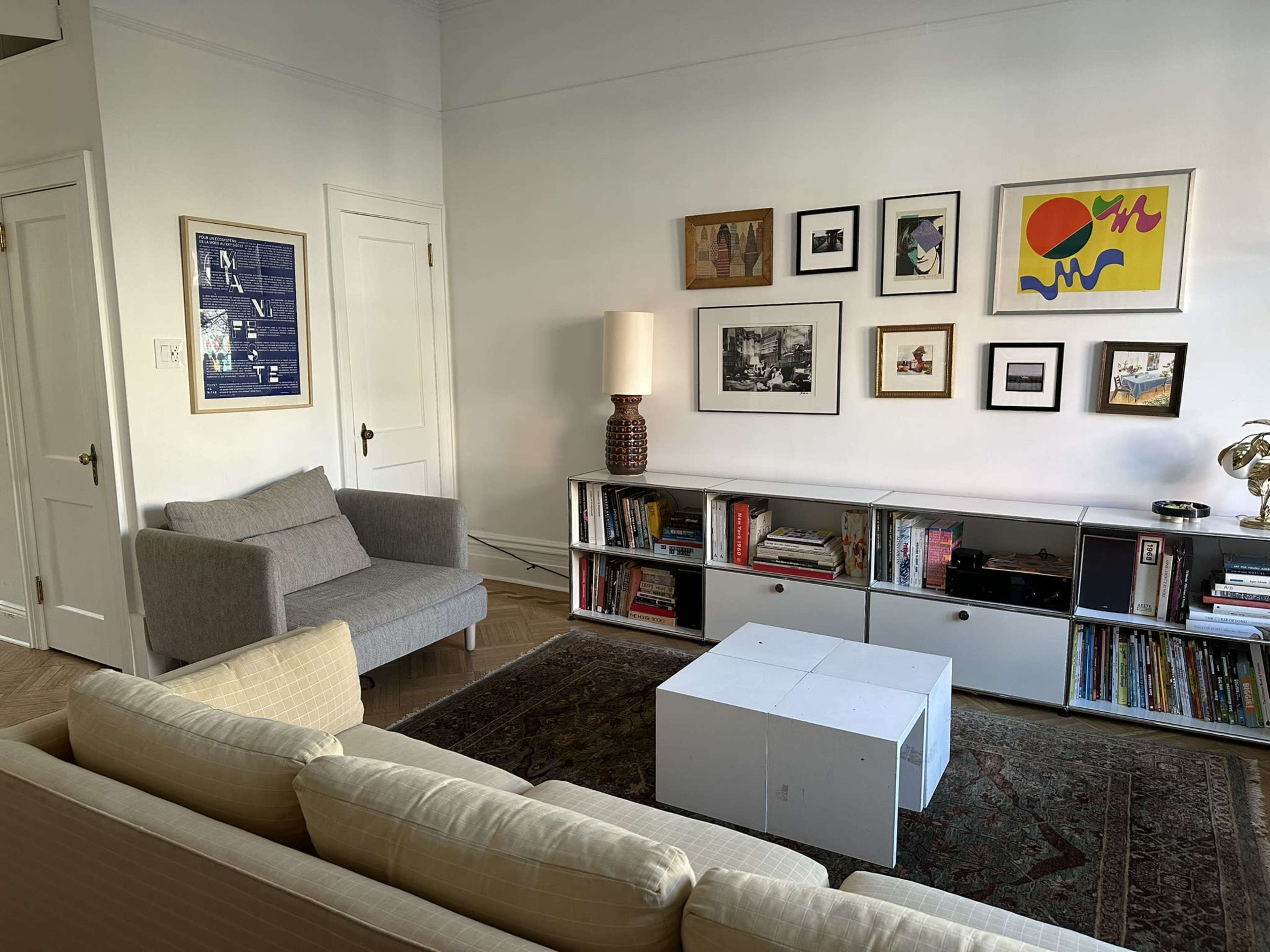 The image shows a well-lit living room featuring a sofa, an armchair, a coffee table, and a media console with books and decorative items against a wall adorned with framed artwork.