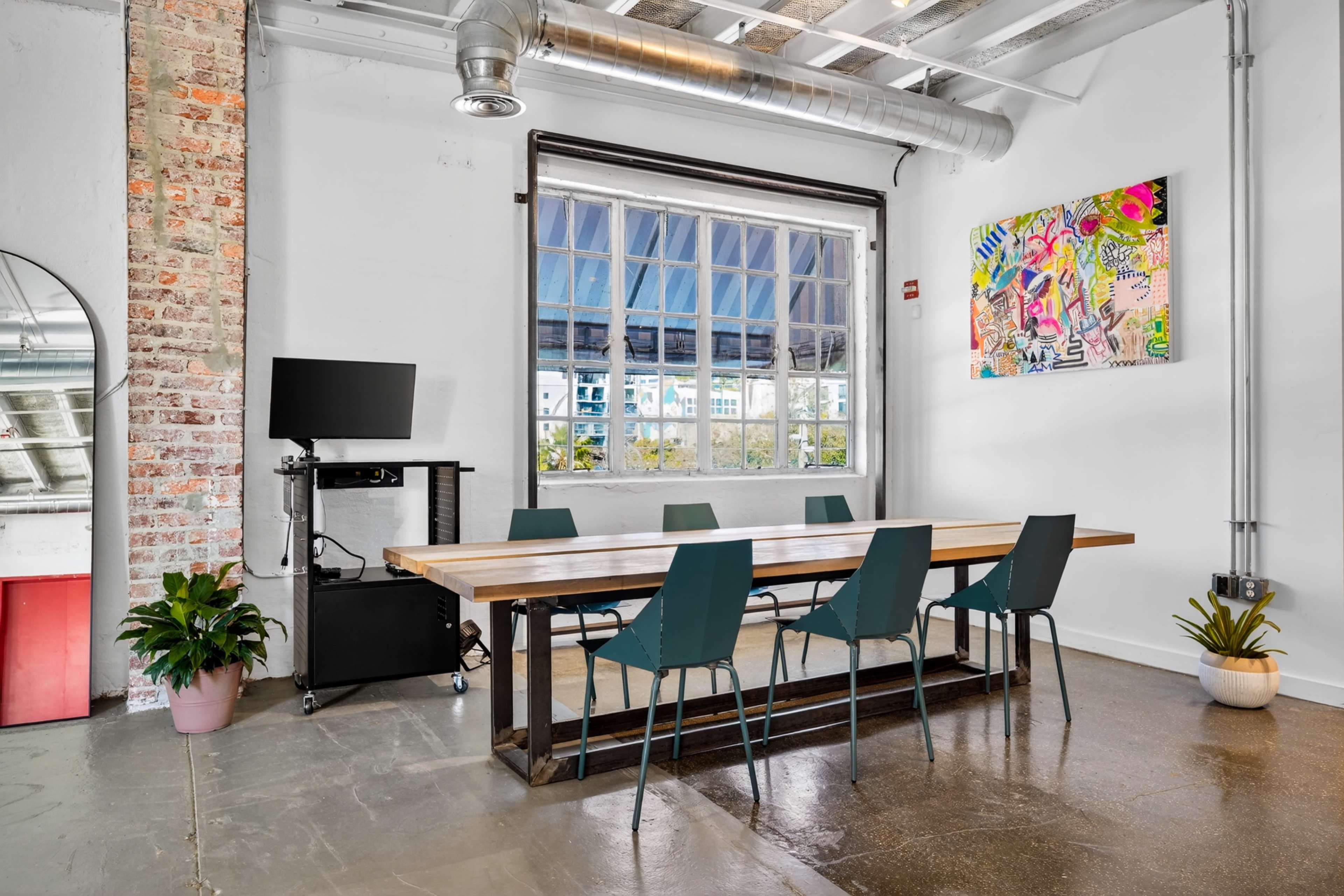 A conference room with a long wooden table surrounded by teal chairs, featuring a large window, a television on a cart, and a colorful artwork on the wall.