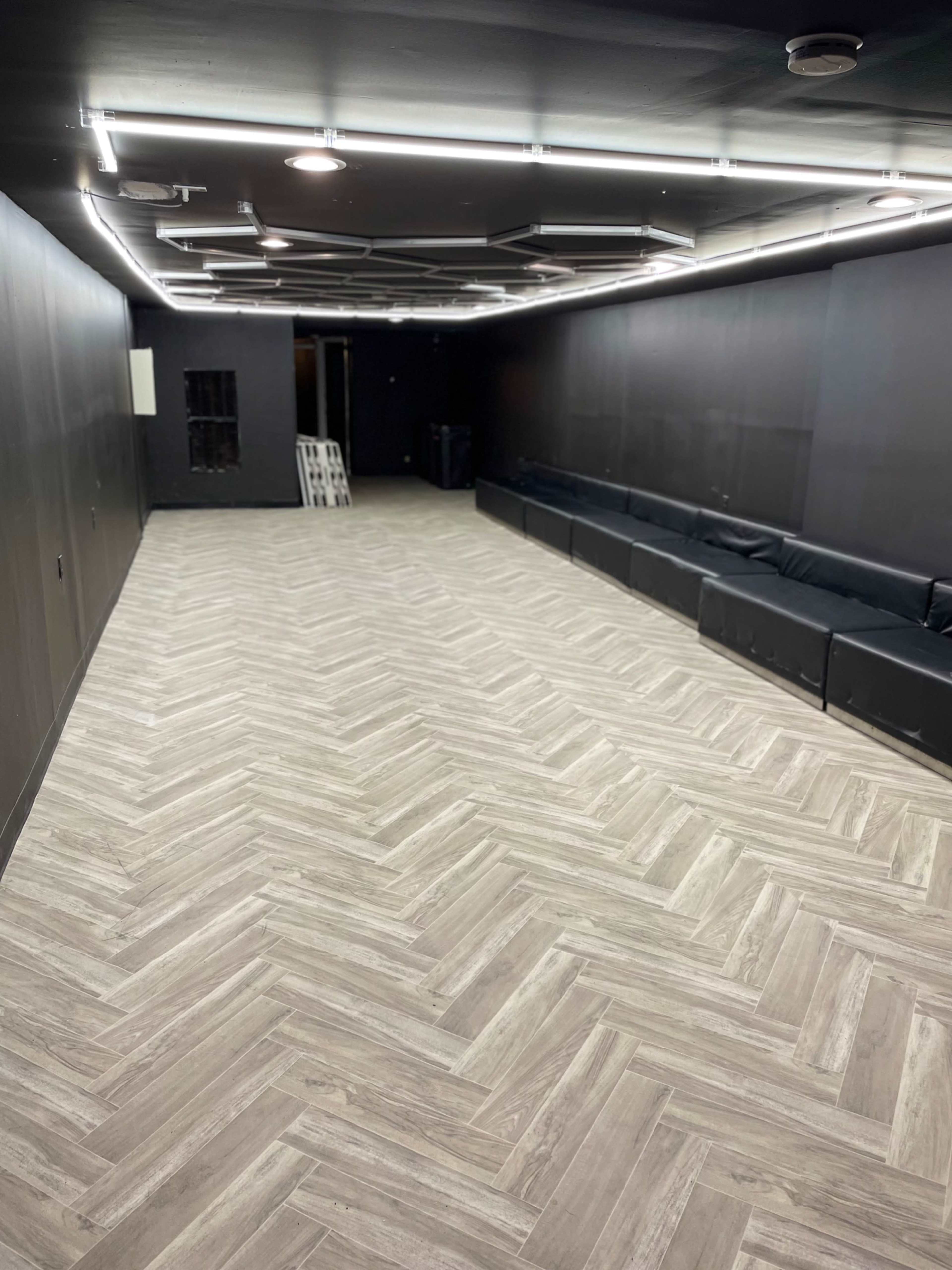 A dimly lit room with herringbone-patterned flooring, black walls, and a long black couch along one side.