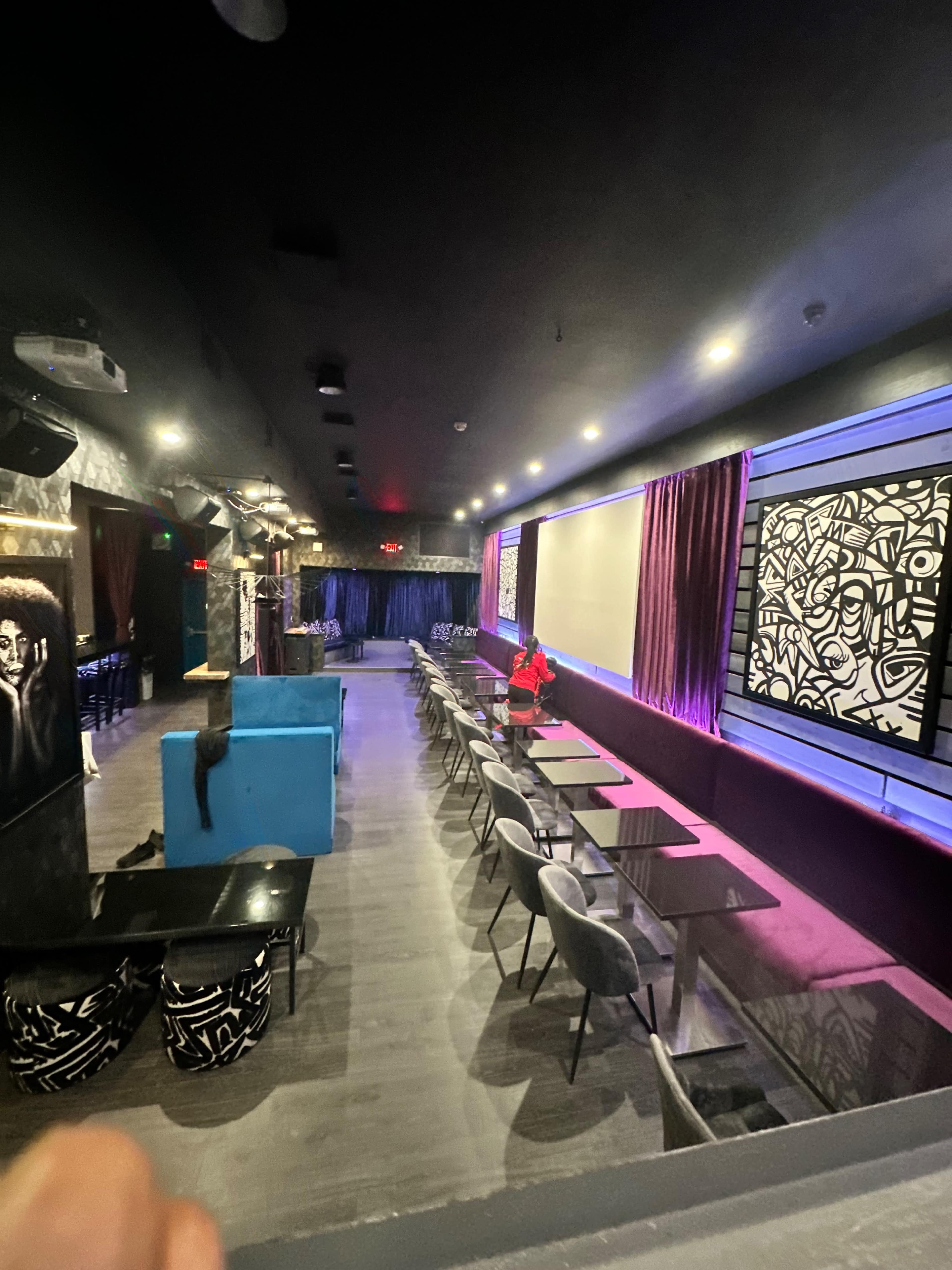 A dimly lit lounge features rows of tables and chairs along the walls, with vibrant artwork and a central stage area.