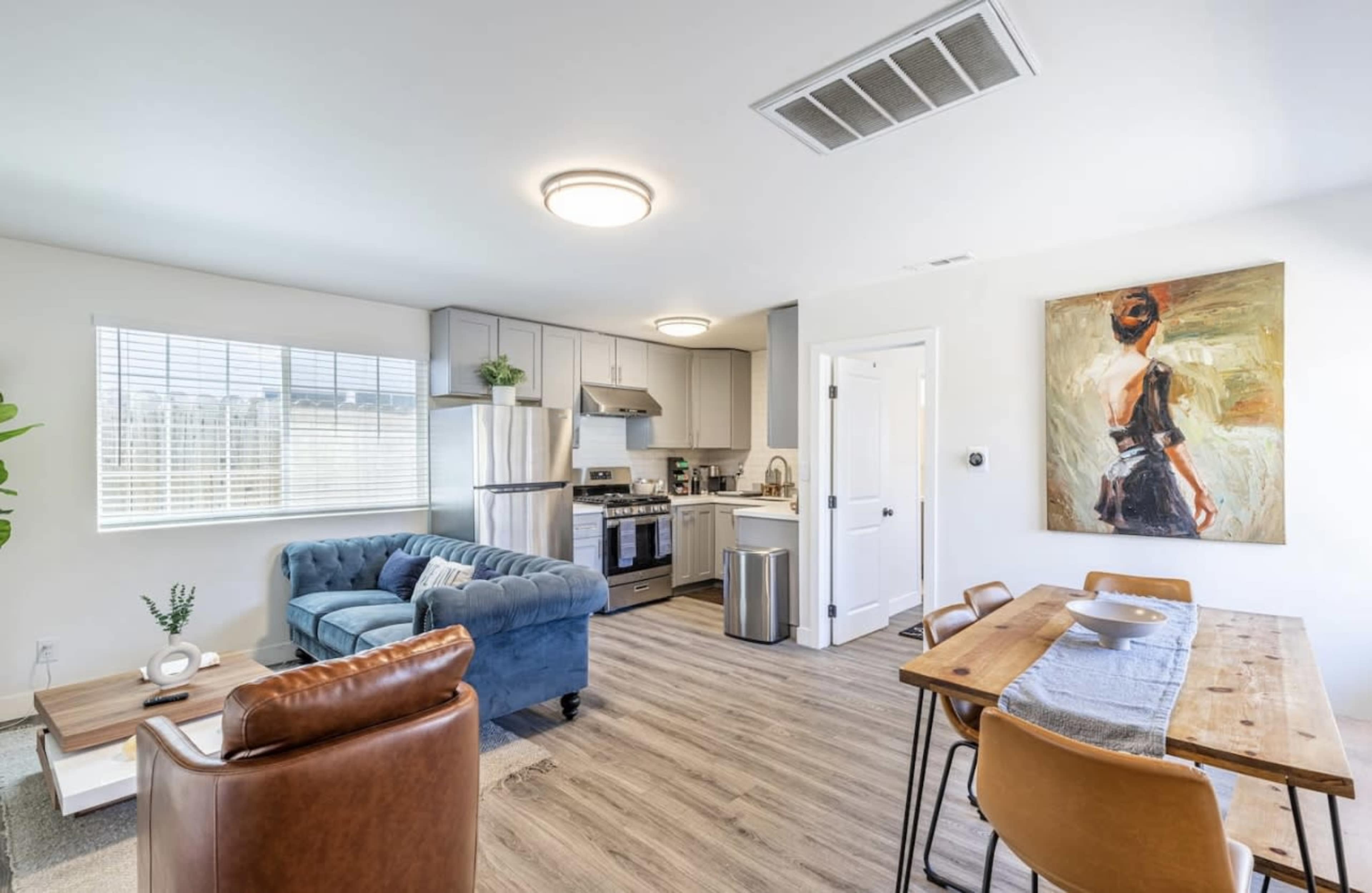 A modern living space features a blue velvet couch, a wooden dining table, and a kitchen with stainless steel appliances.