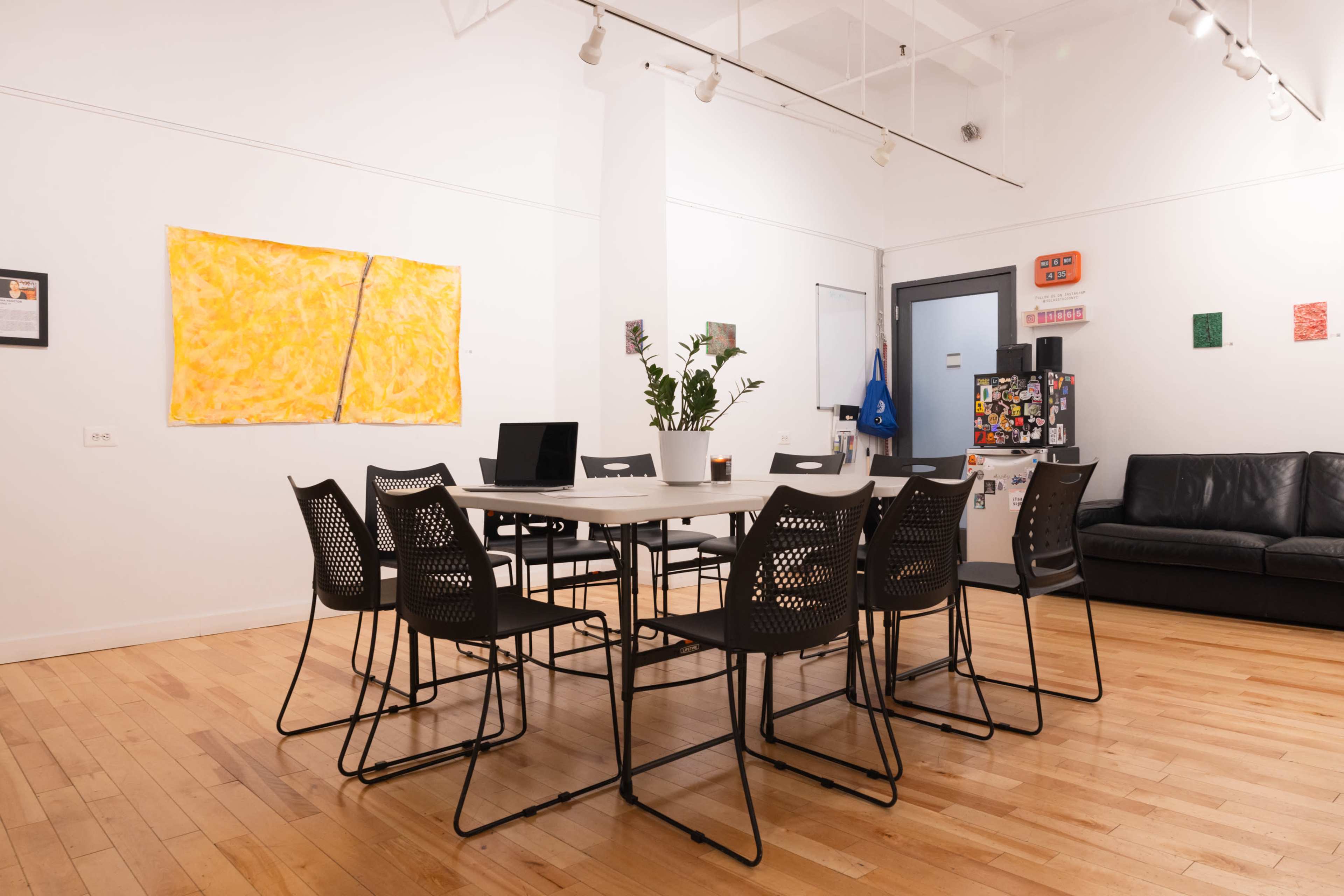 The image shows a modern meeting room with a large table surrounded by black mesh chairs, a potted plant in the center, and various artworks displayed on the walls.