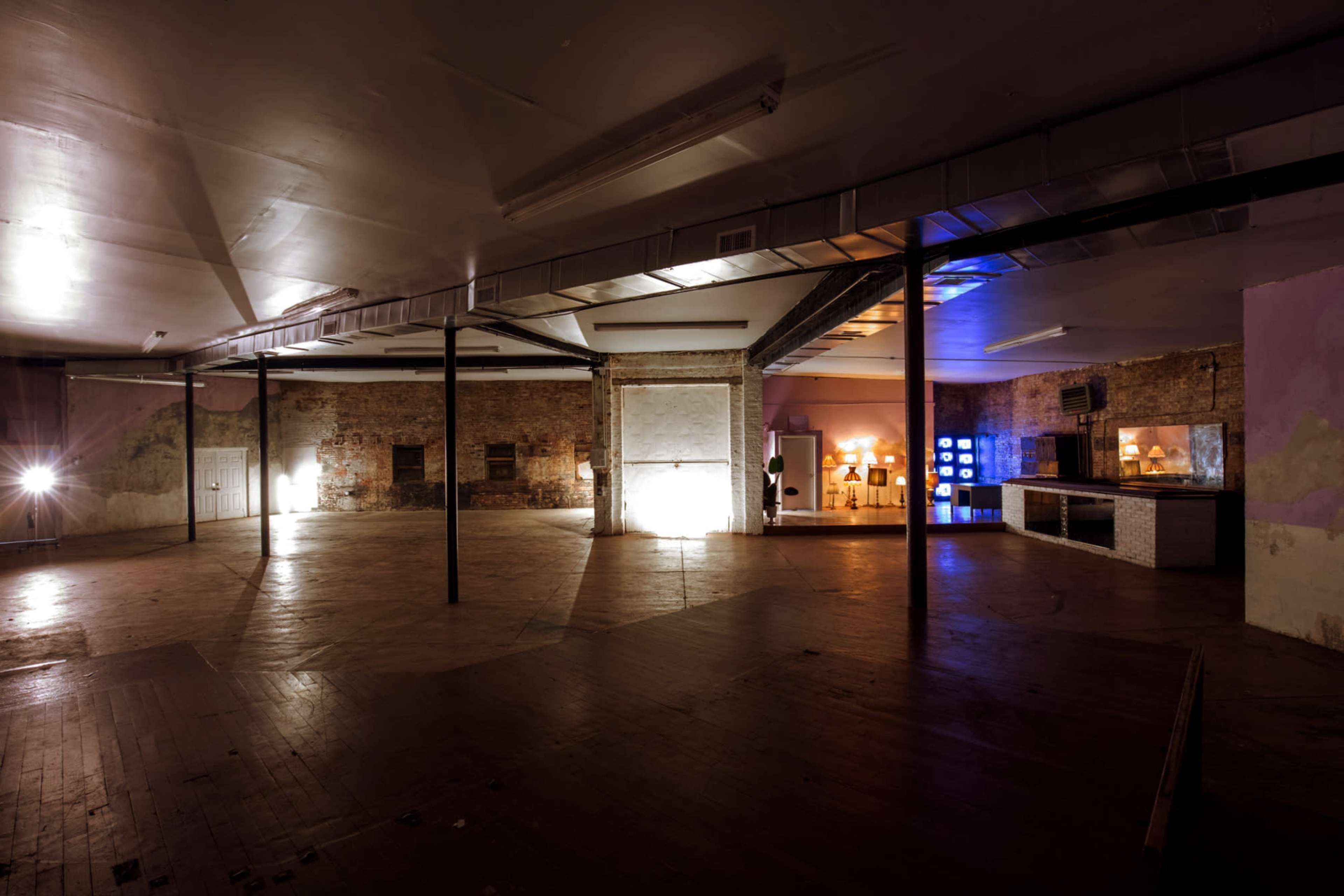The image shows an empty, spacious interior of a building with exposed brick walls and wooden flooring, featuring sparse lighting and a few decorative elements in the background.