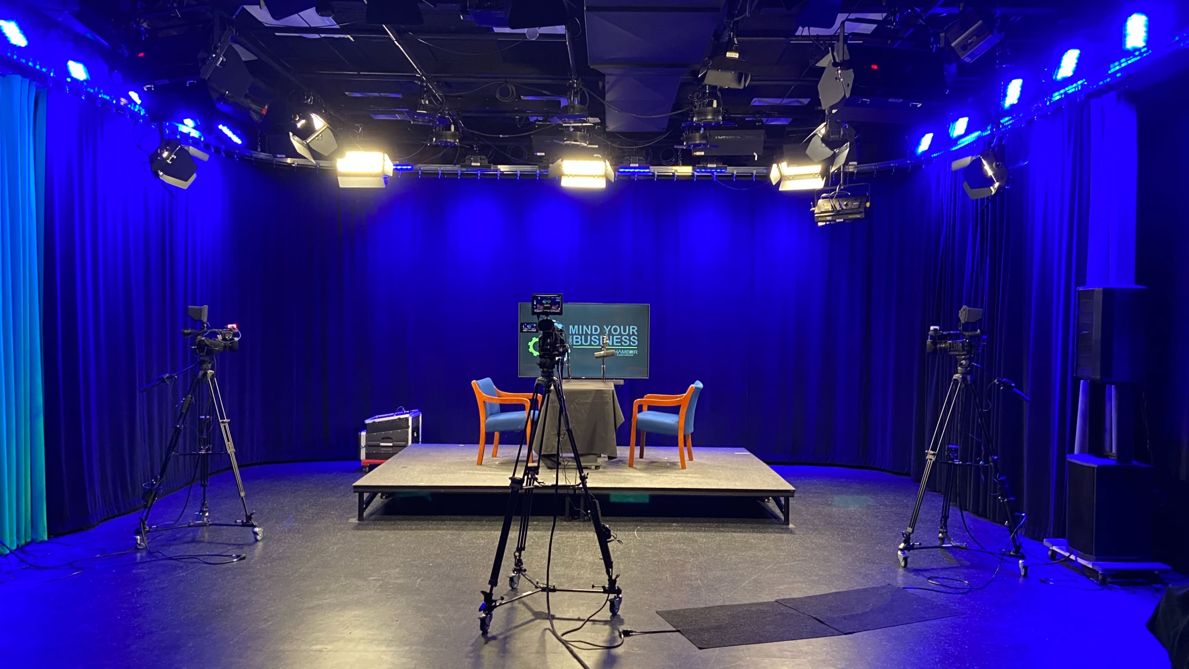 A television studio is set up with cameras facing a central table and two chairs, illuminated by various lights against a dark backdrop.