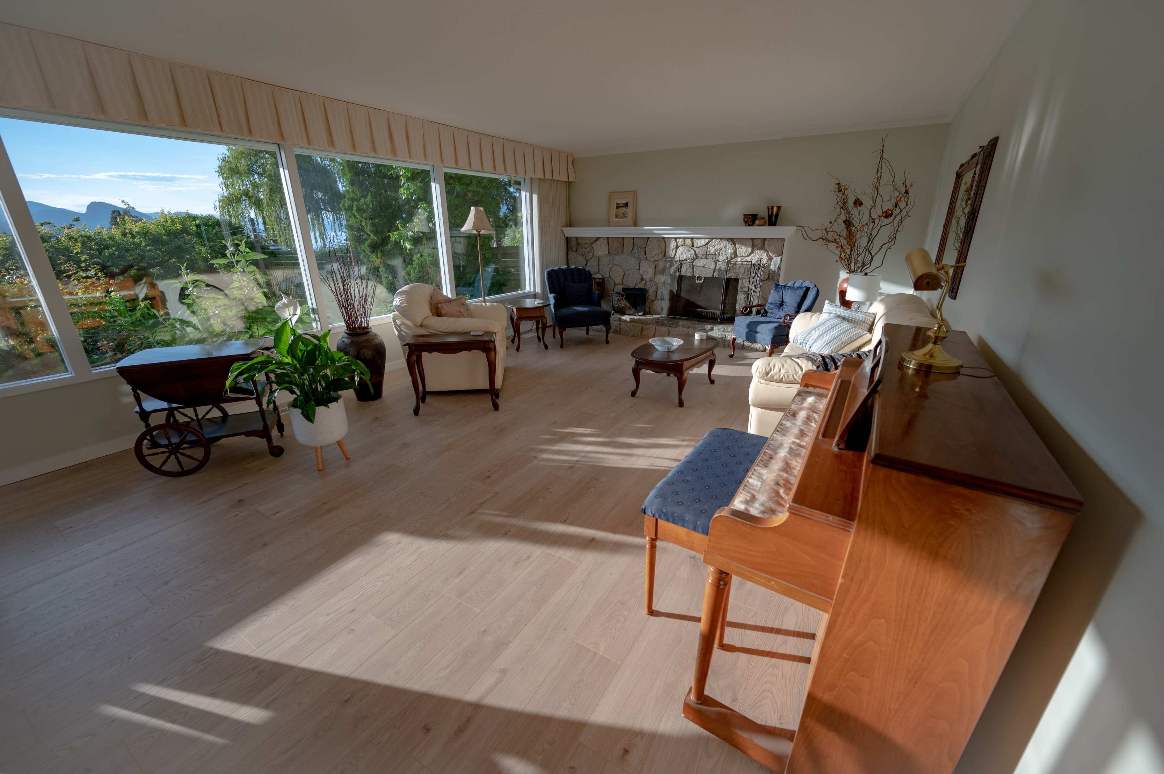The living room features large windows, a stone fireplace, and a mix of seating arrangements including chairs and a sofa, with a piano and decorative plants in the space.