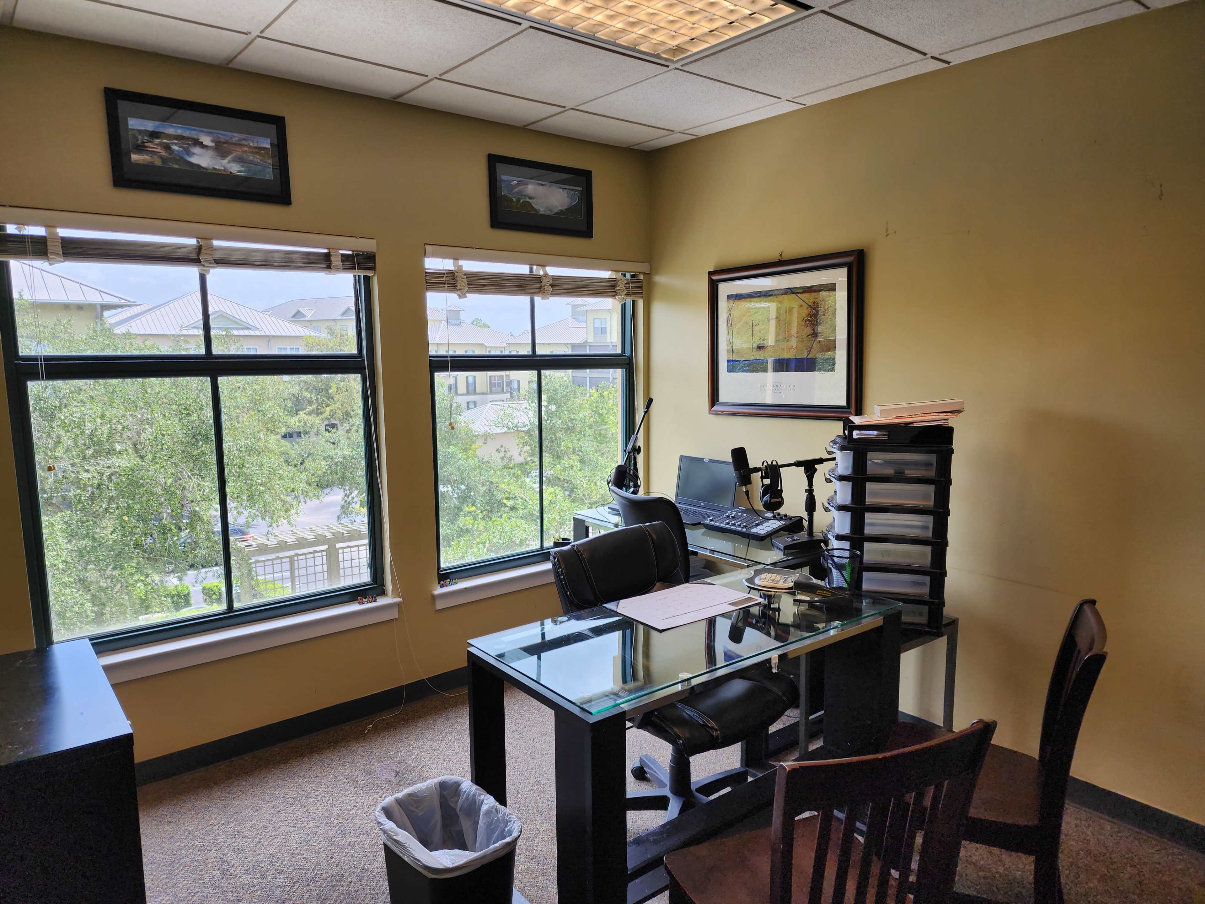 The image shows an office with a glass-top desk, a chair, and a computer setup near large windows overlooking trees and buildings.