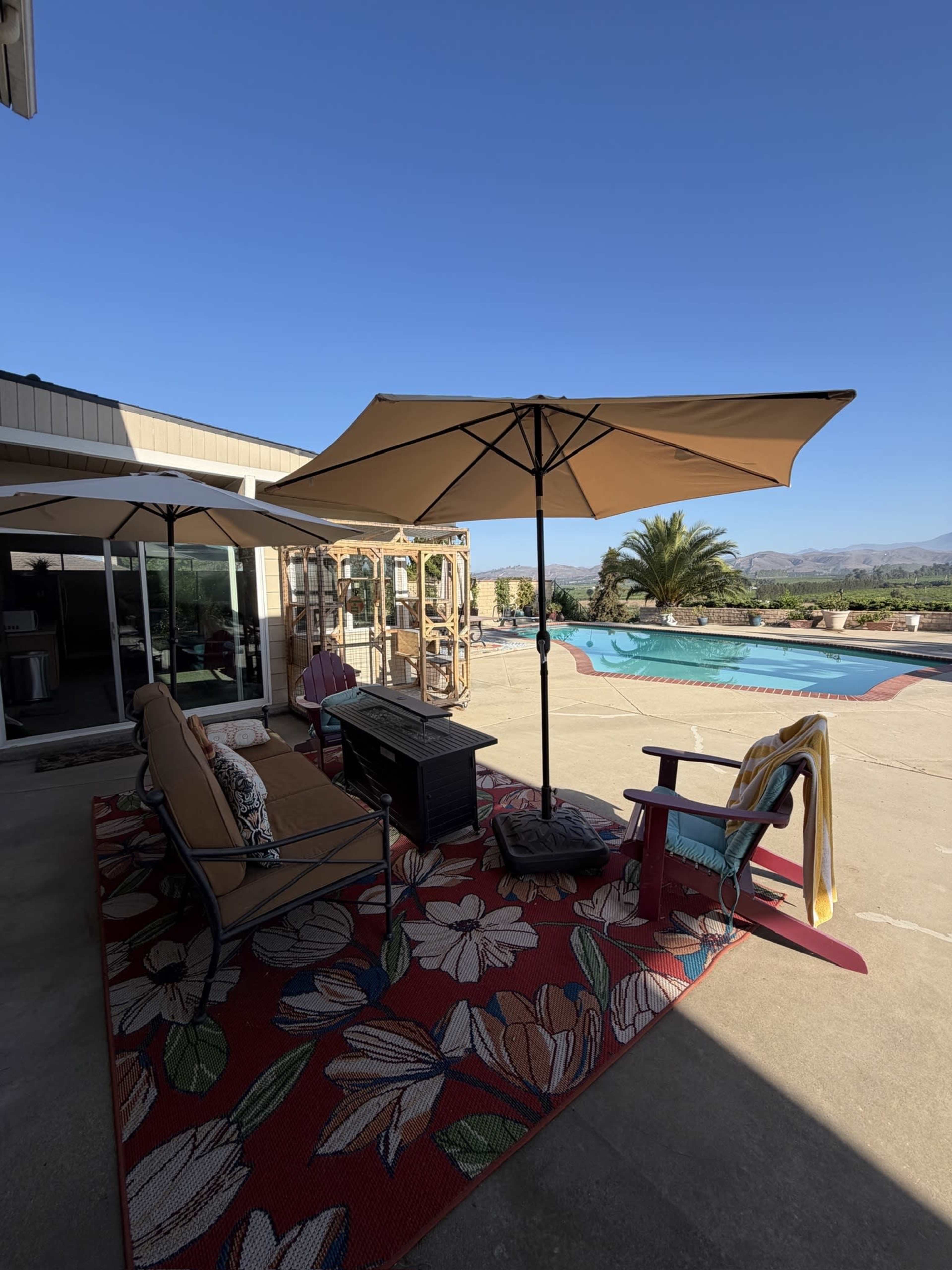 The image shows a poolside seating area with two umbrella-covered chairs, a small table, and a view of a swimming pool and mountains in the background.