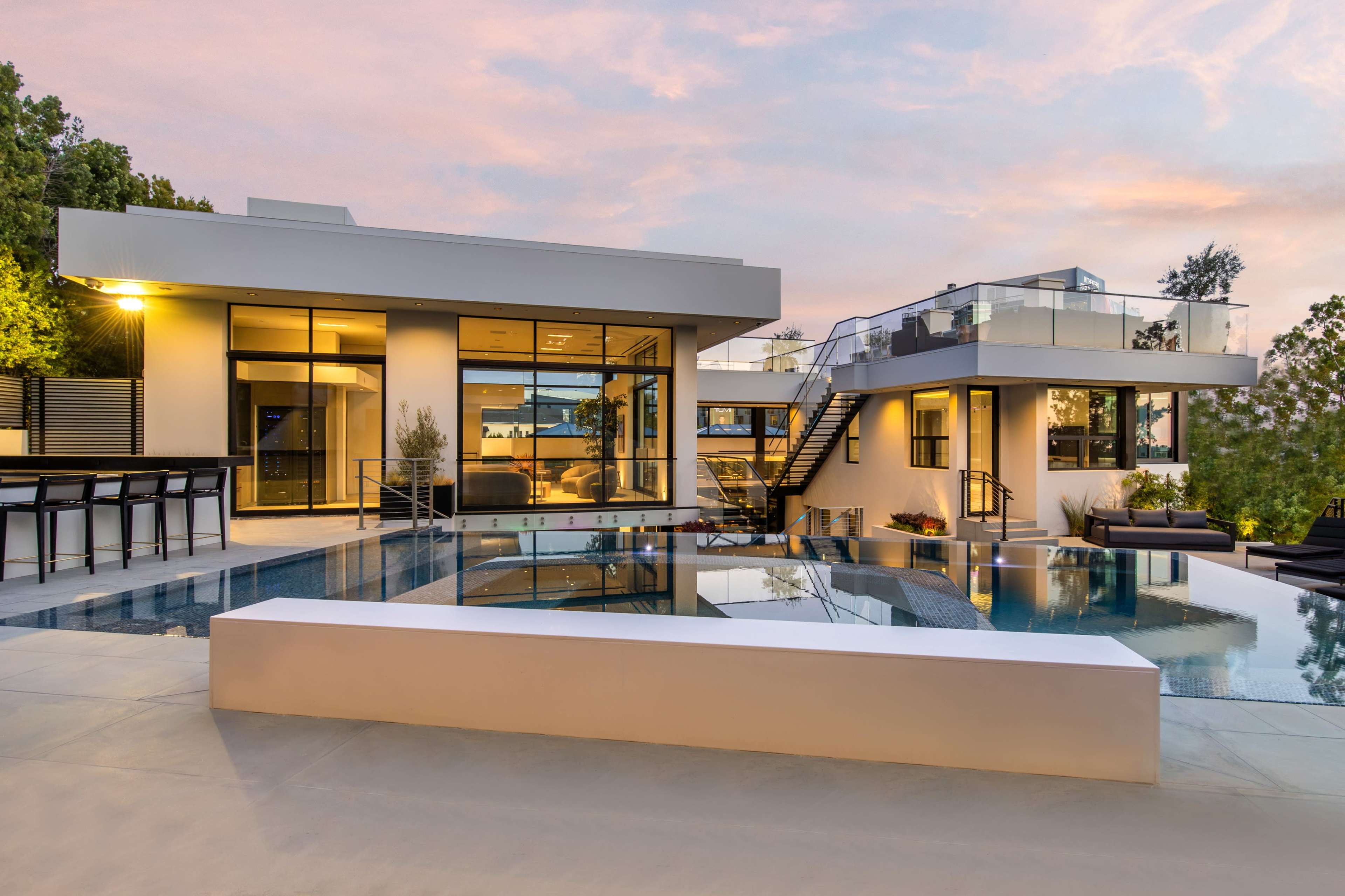 The image shows a modern two-story house with large glass windows overlooking a swimming pool and outdoor patio area, surrounded by greenery and a sunset sky.