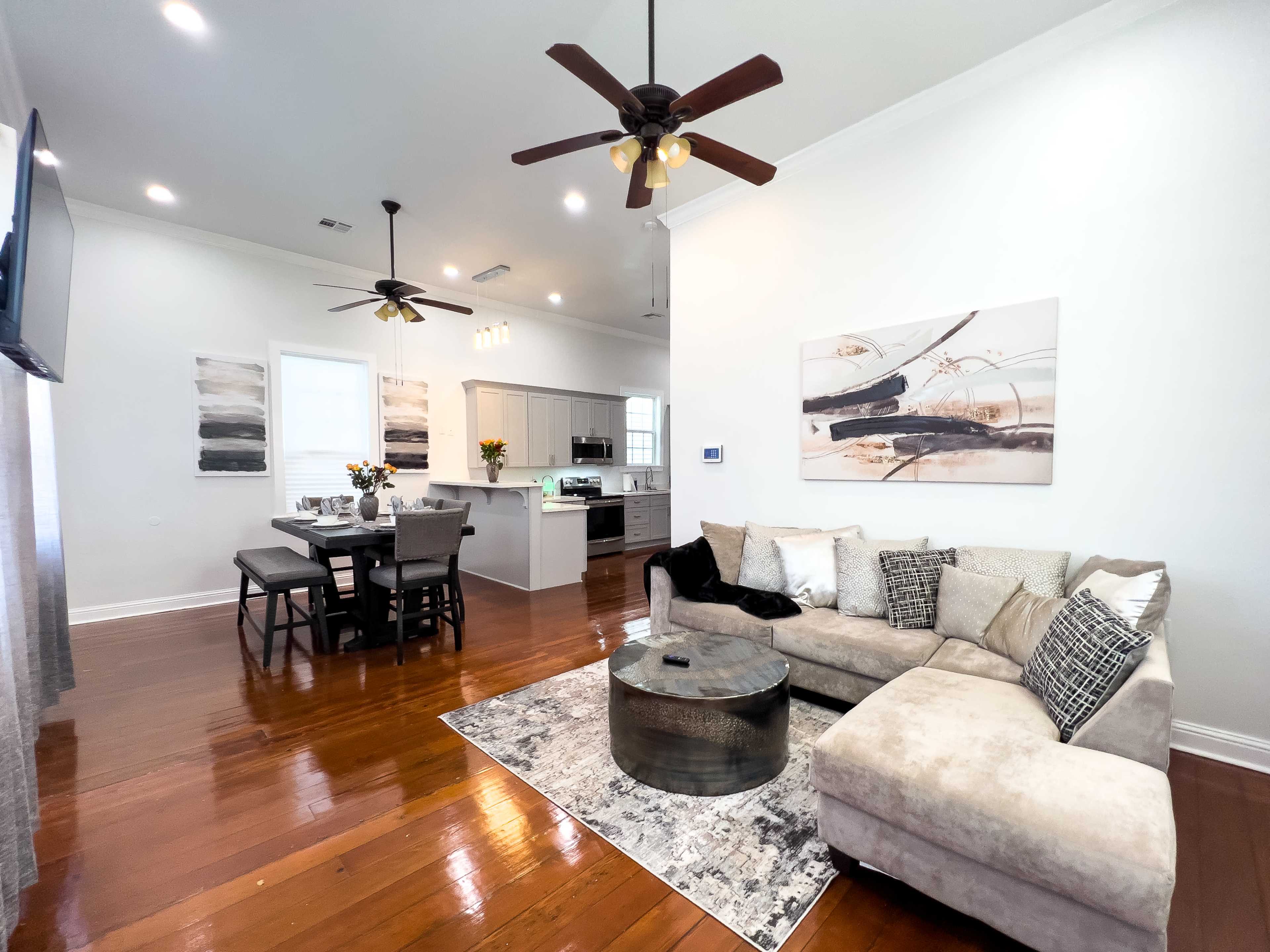 The image shows a modern living room and dining area with hardwood floors, featuring a sectional couch, a circular coffee table, and a dining table set for four.