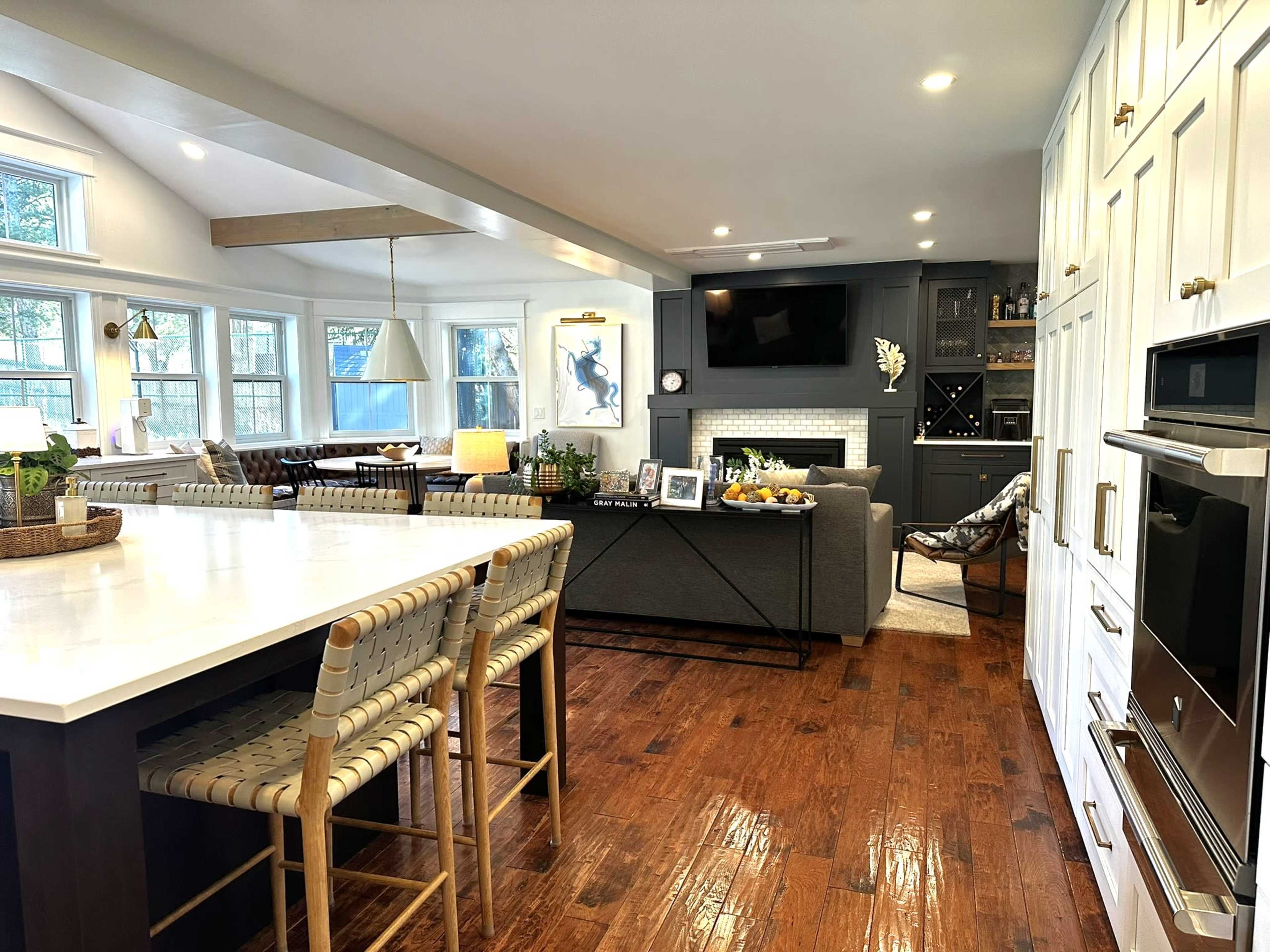 A modern kitchen with a large island and bar stools opens up to a cozy living area featuring a gray sofa and a fireplace.