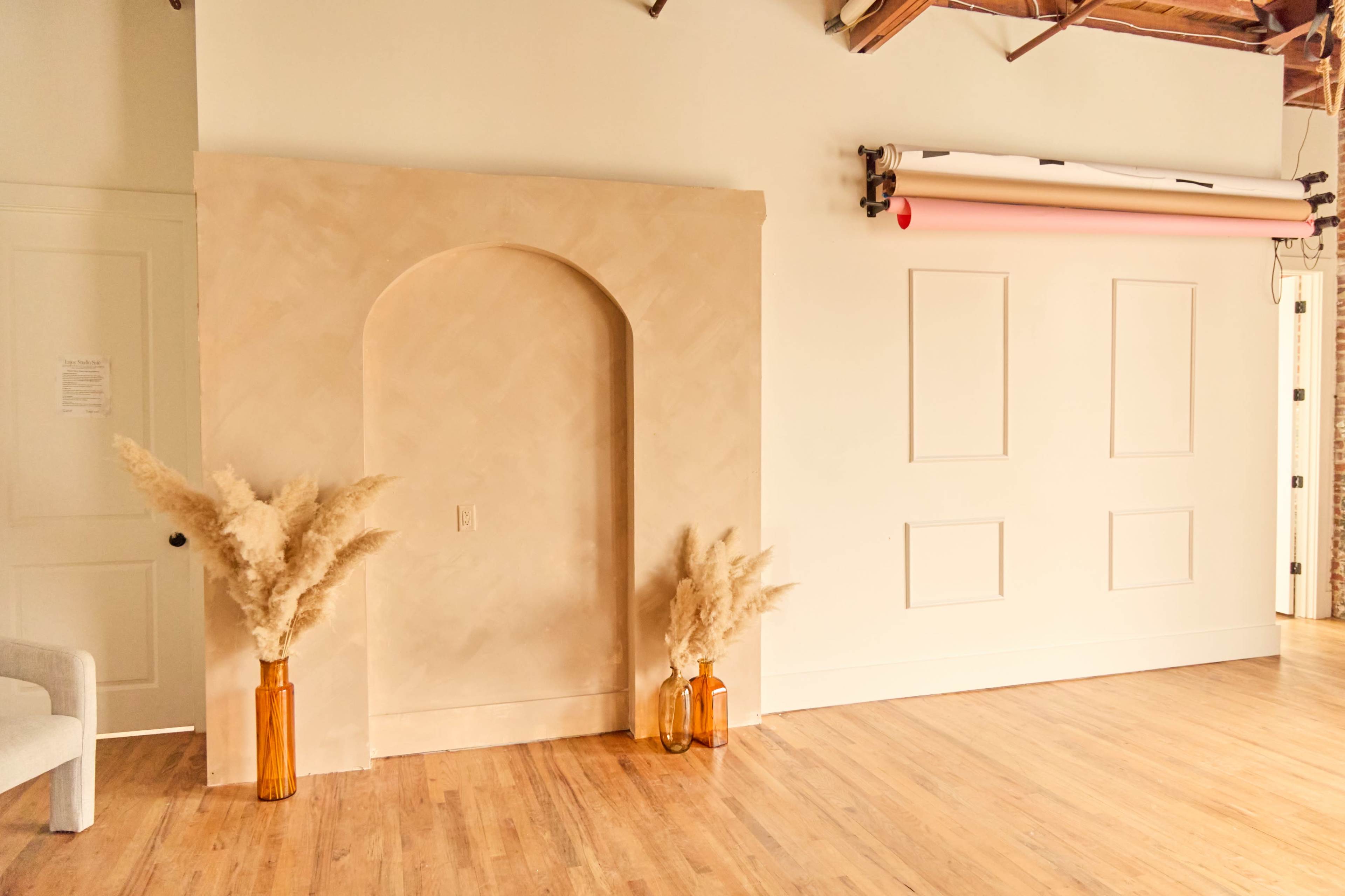 A room features a smooth archway on one wall, flanked by two vases holding pampas grass, with a backdrop of neutral-colored walls and wooden flooring.