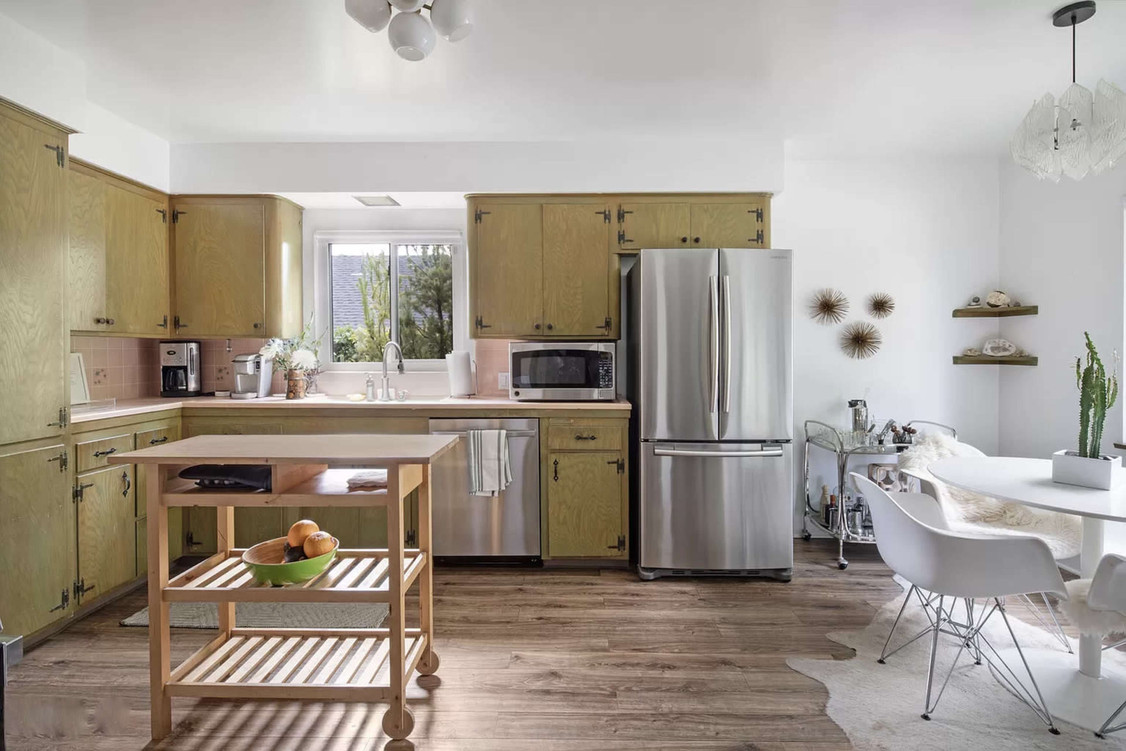 A bright kitchen features wood cabinets, stainless steel appliances, and a dining table with white chairs beside a countertop on wheels.