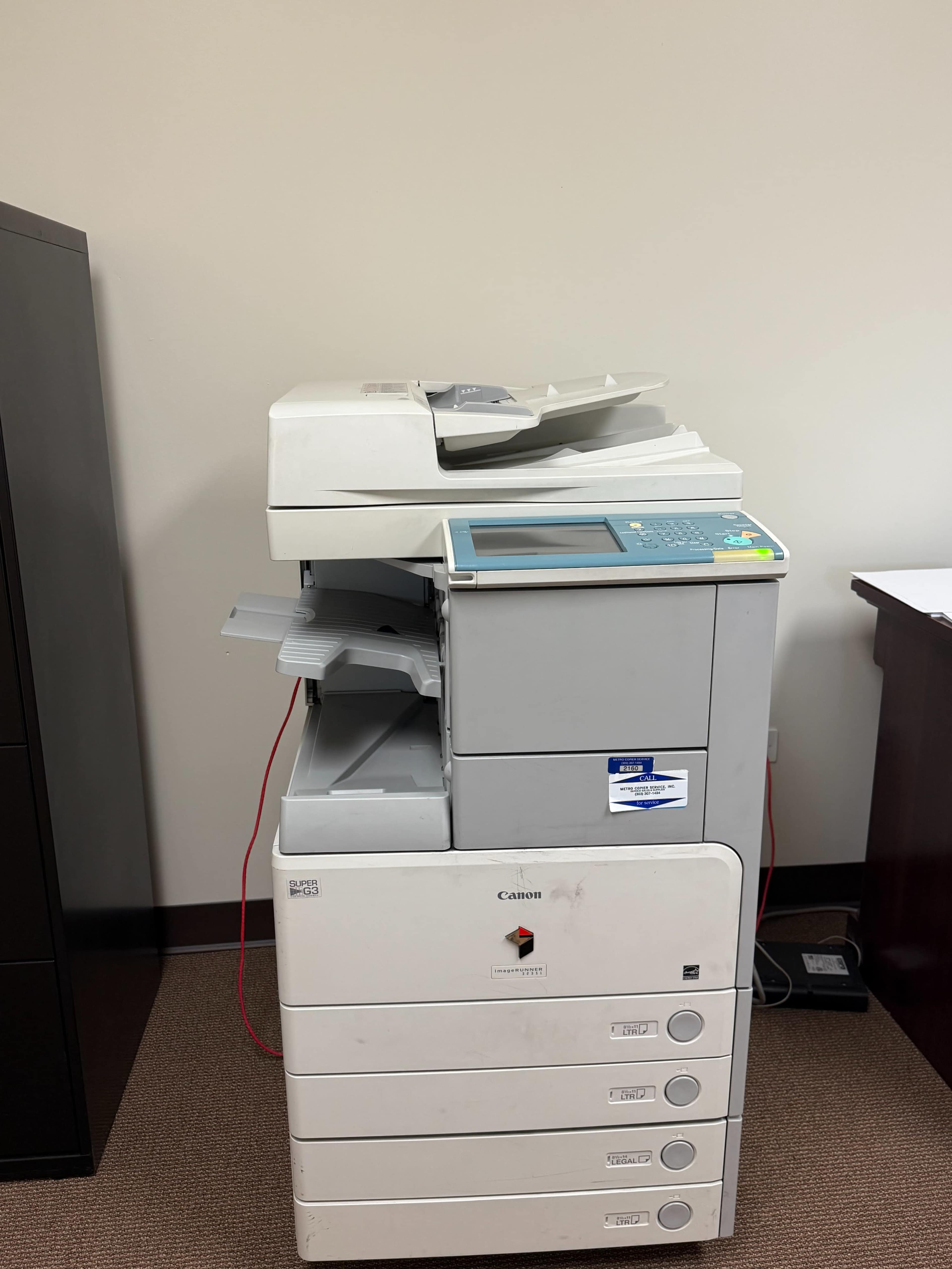 A monochrome Canon copier with multiple paper trays is positioned against a beige wall in an office setting.