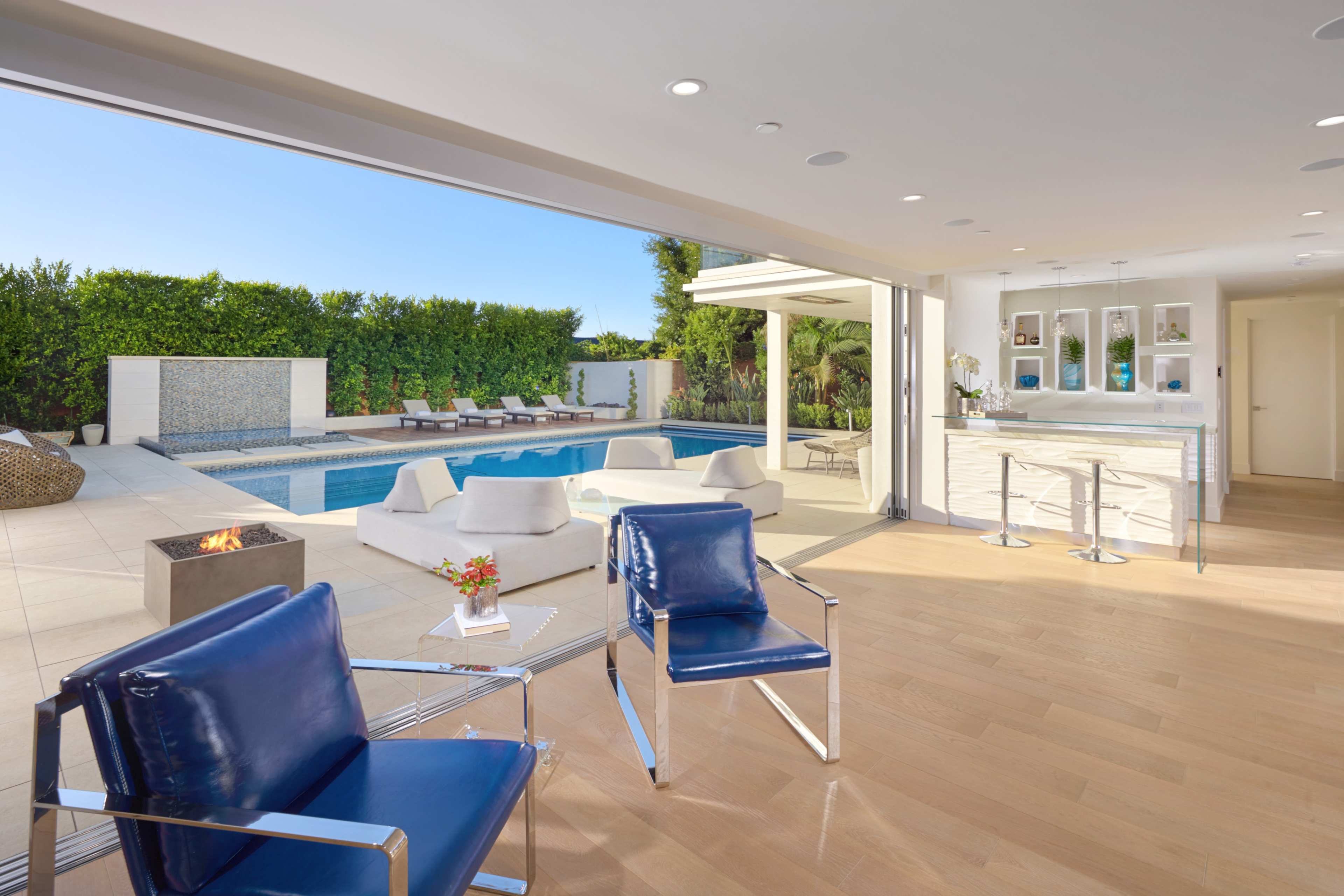 A modern indoor space with blue chairs, opening to a patio with a swimming pool and sun loungers surrounded by greenery.