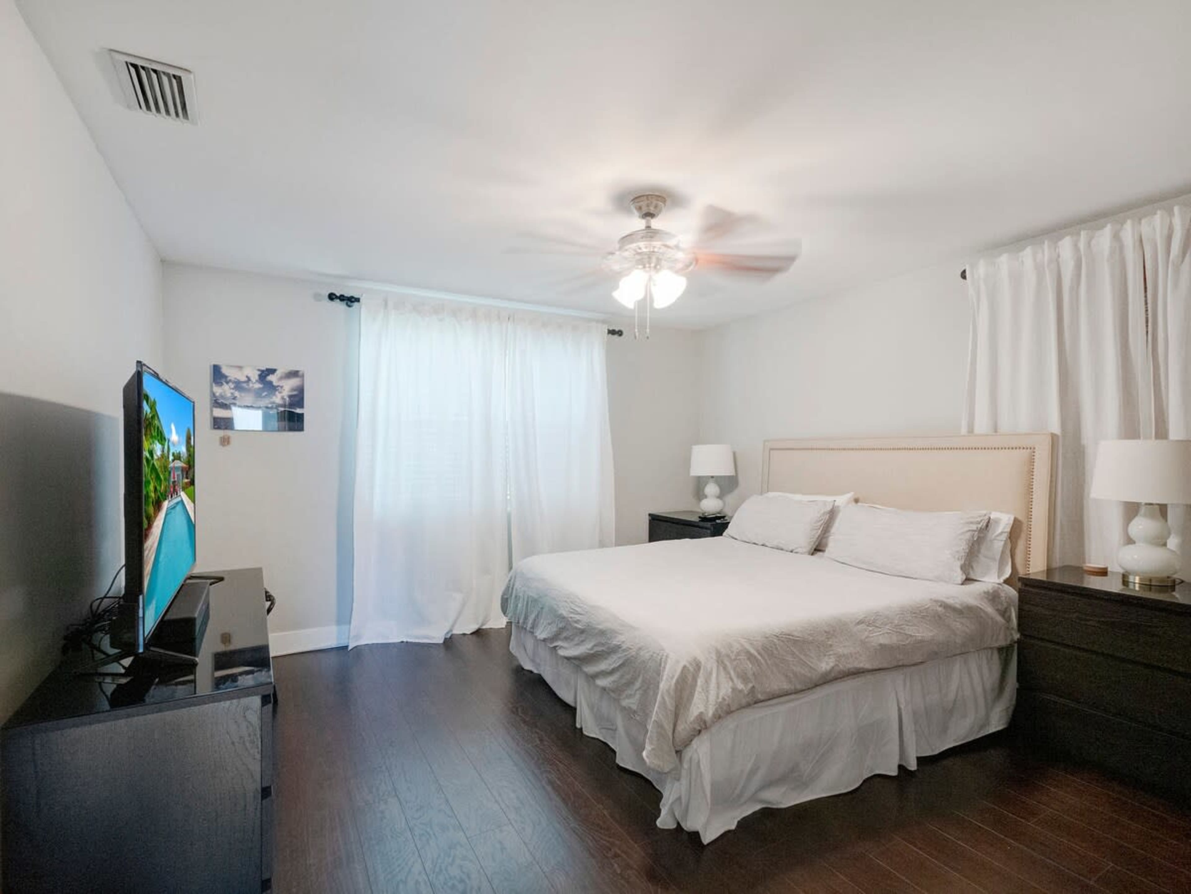 A spacious bedroom features a light-colored bed, a television on a dark dresser, and large windows covered with white curtains.