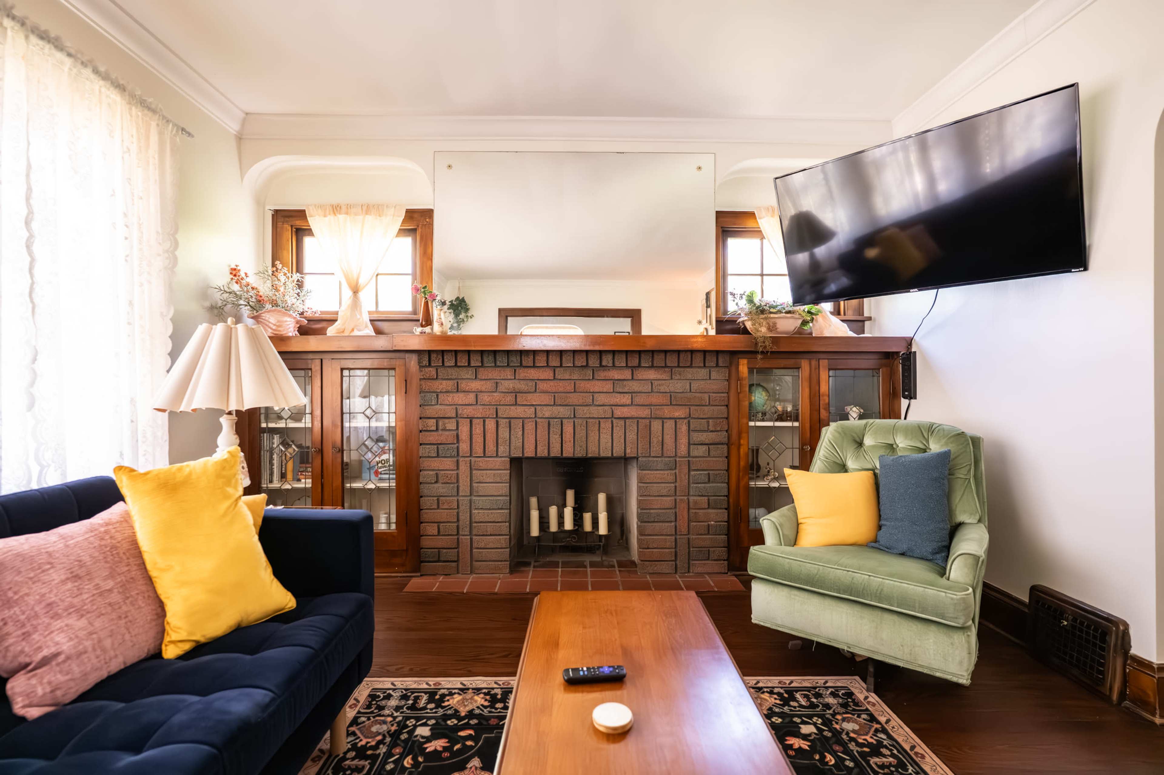 The image shows a living room with a blue sofa, a green armchair, a brick fireplace, and a mounted television.
