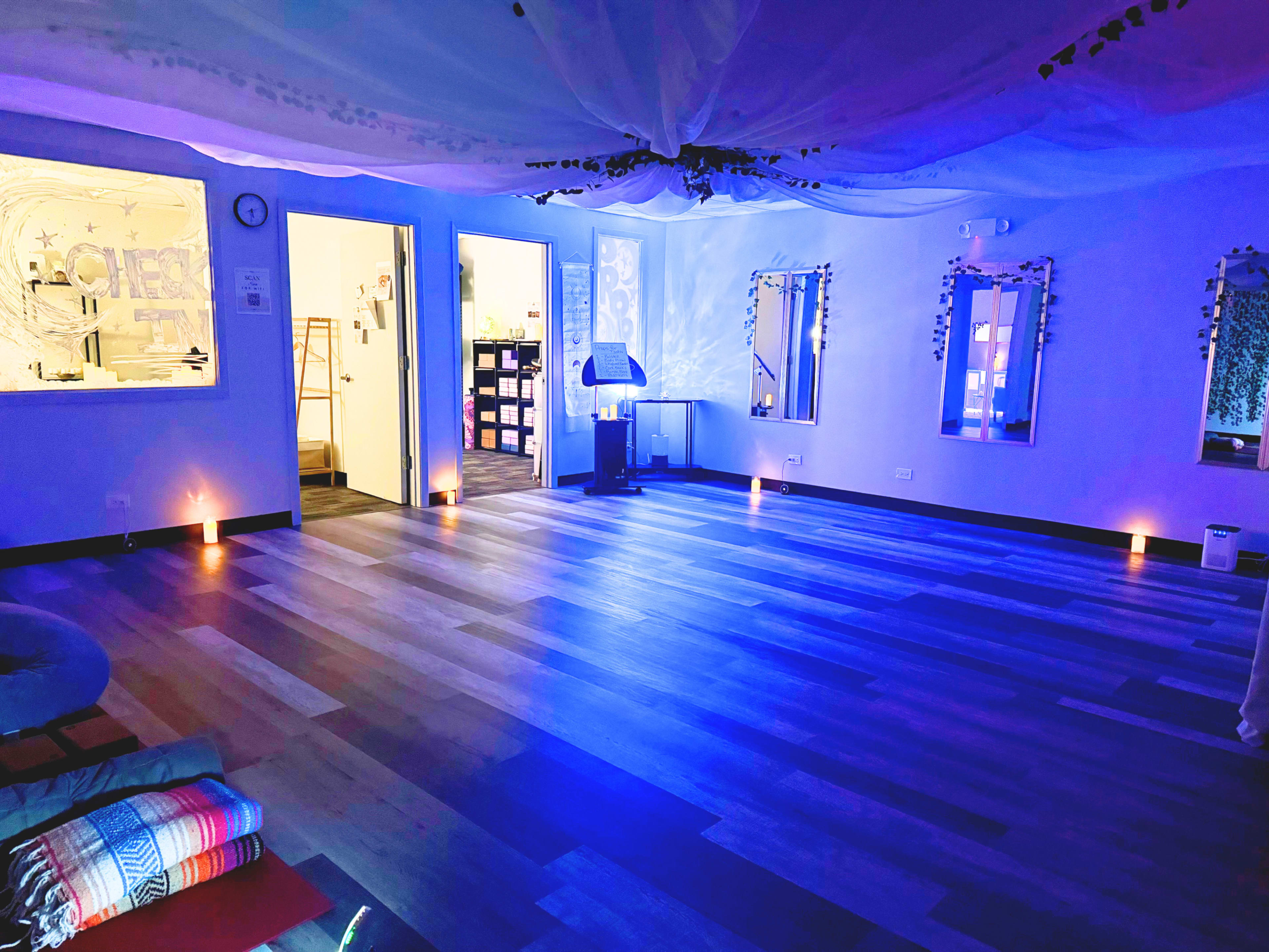 The image shows a spacious, softly lit room with wooden flooring, mirrors on the walls, and a calming blue hue from the lighting.