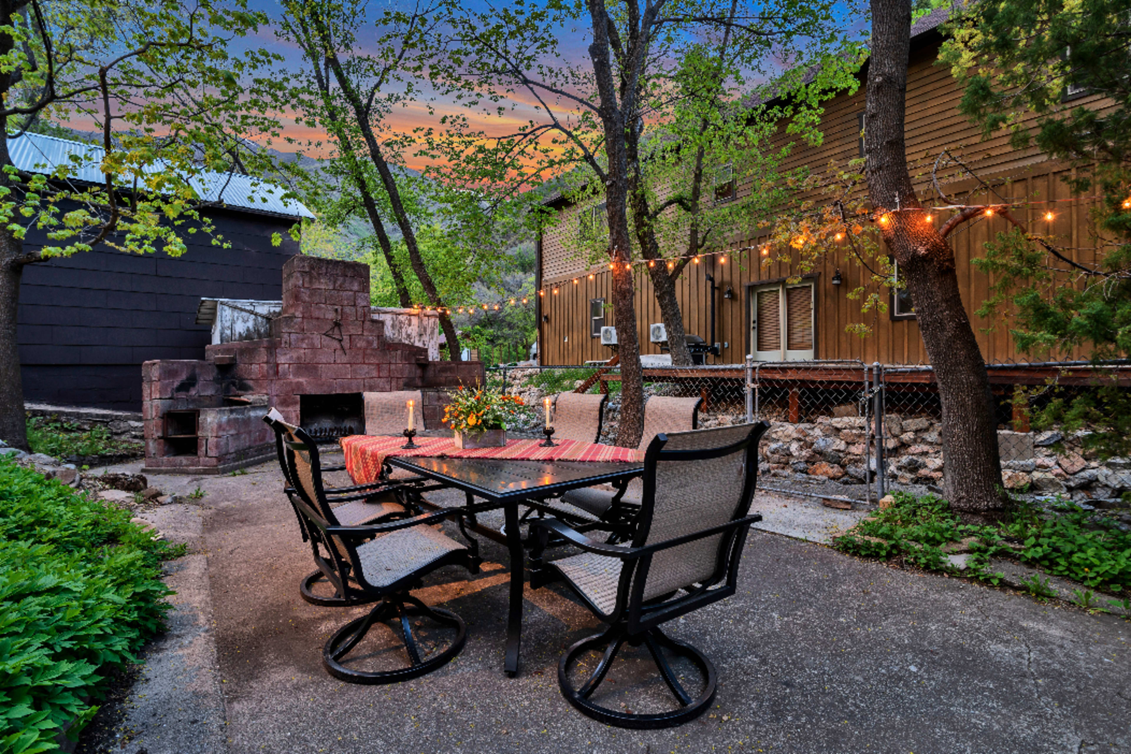 A patio area featuring a dining table with chairs, an outdoor fireplace, and decorative string lights, surrounded by trees and buildings.