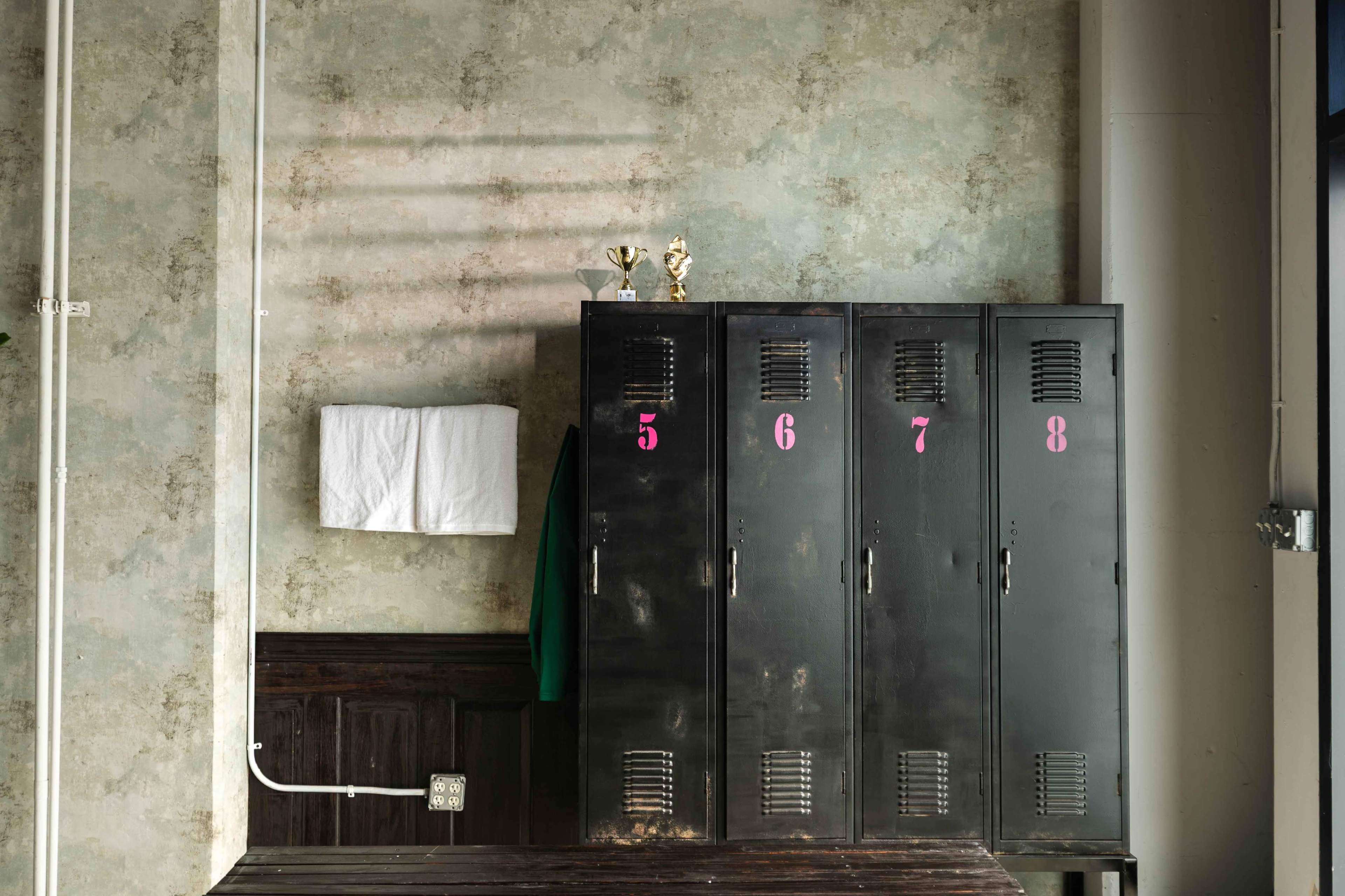 The image shows a set of black lockers with numbers 5, 6, 7, and 8, mounted on a weathered wall, alongside a towel hanging and trophies on top.