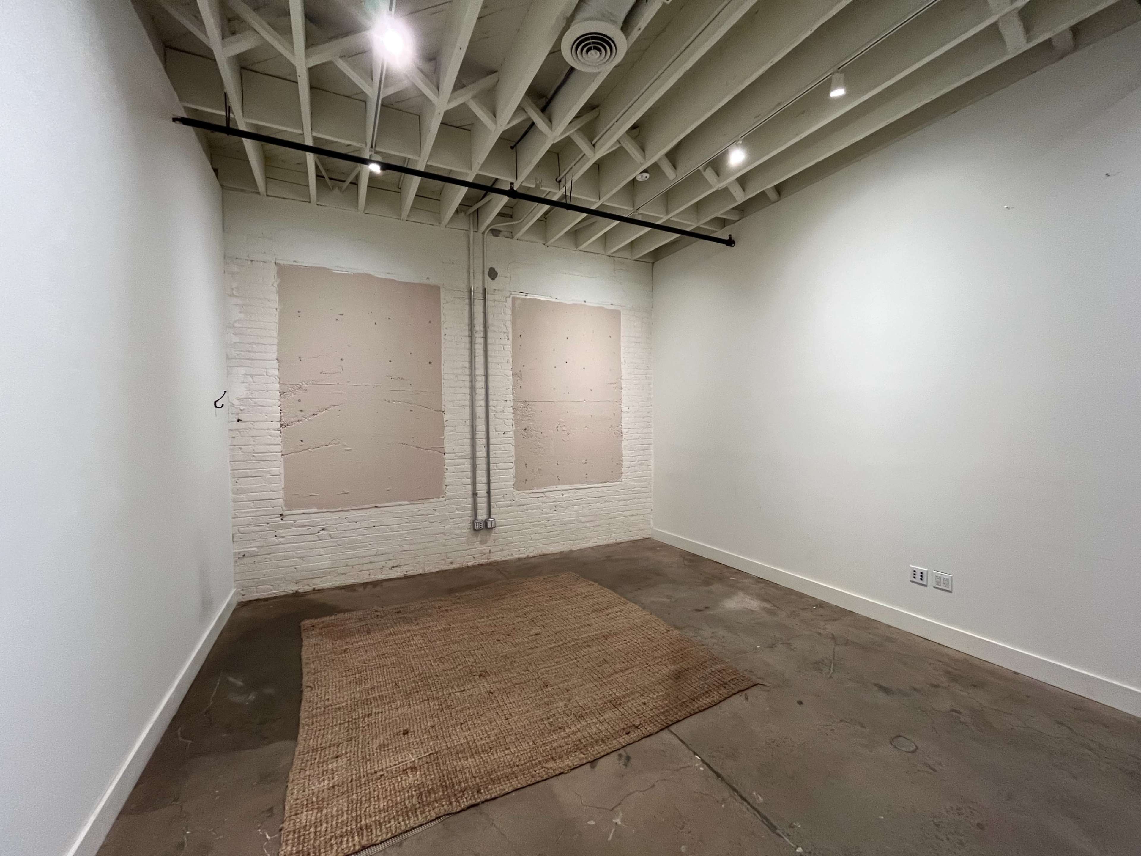A minimalist, empty room with exposed beams, two bare wall sections, and a woven rug on the concrete floor.