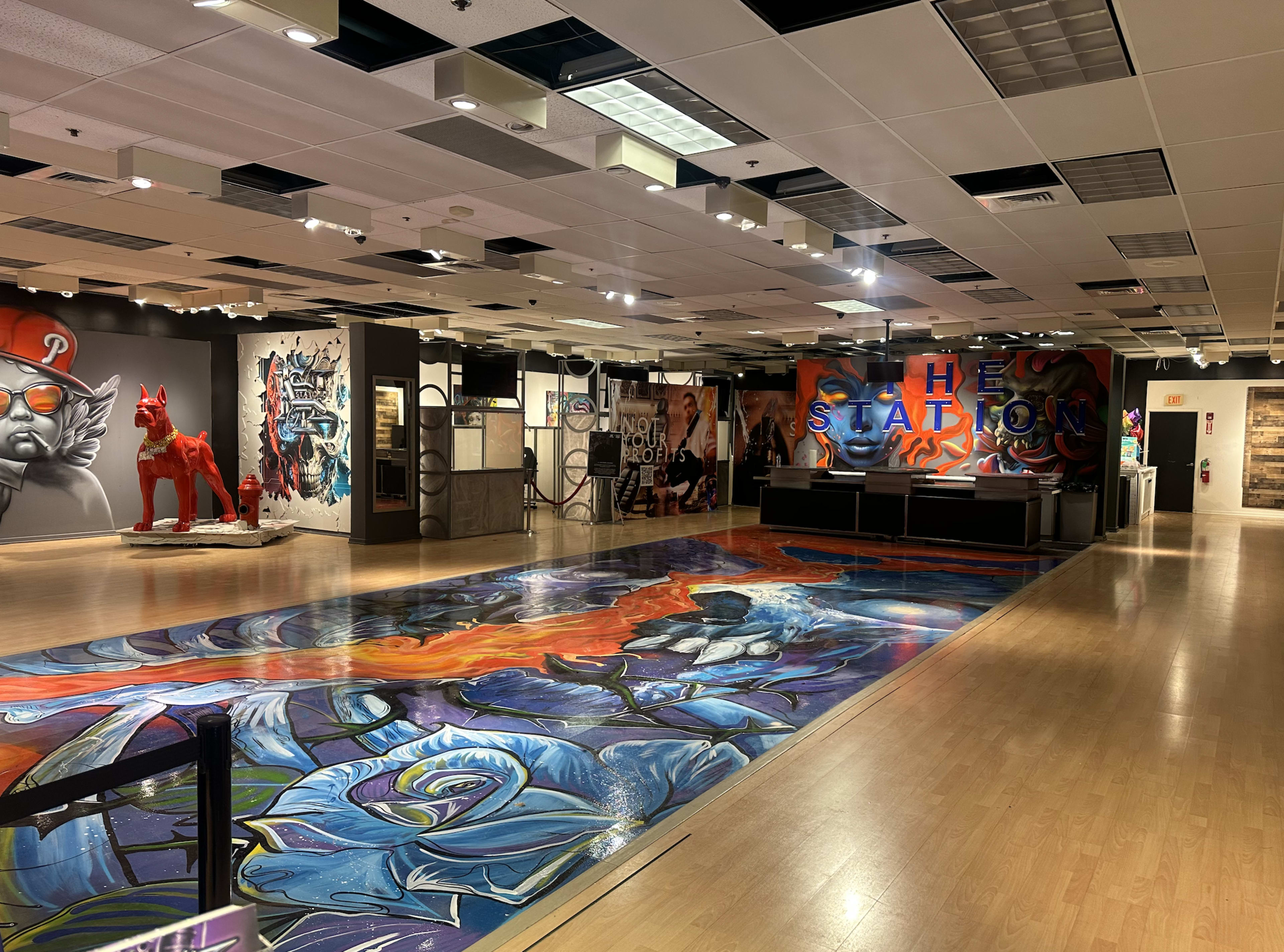 The image shows an art gallery featuring vibrant murals on the walls and a large, colorful mural on the floor, with sculptures and exhibits displayed throughout the space.