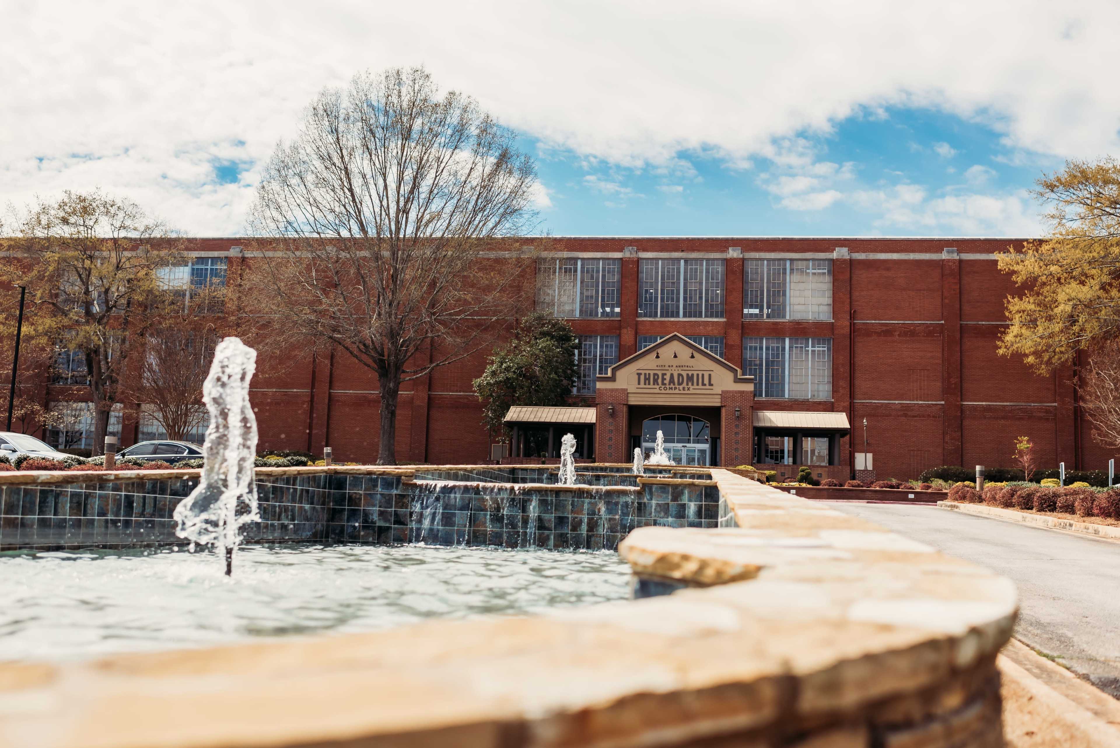 A stone fountain with a small water spout is in the foreground, while a red brick building labeled "THREADMILL" is visible in the background.