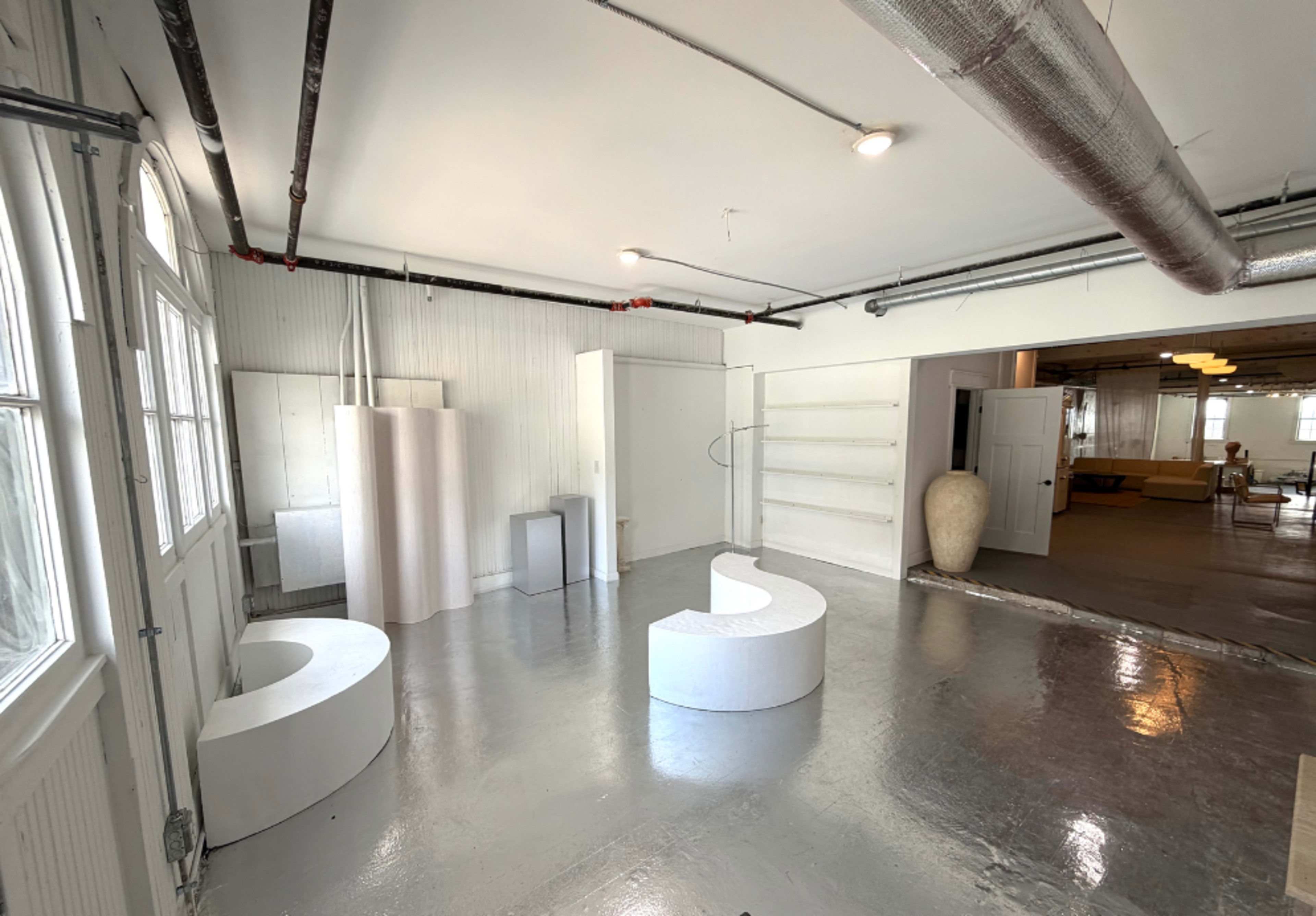 The image shows a spacious, industrial-style room containing minimalist white furniture and fixtures, including curved seating and a tall vase.