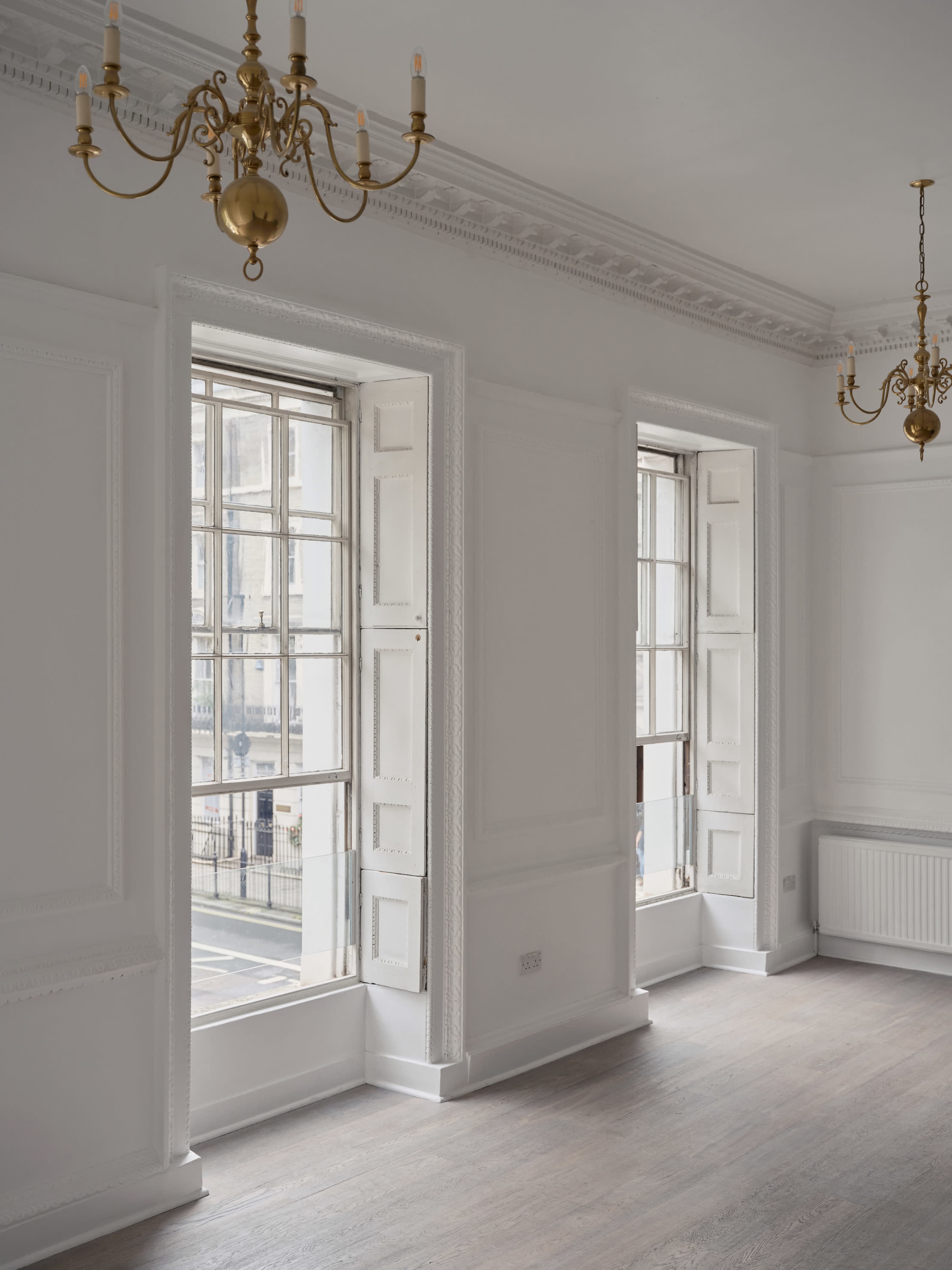 The image shows a bright, empty room with two large windows and ornate chandeliers, featuring white walls and wooden flooring.