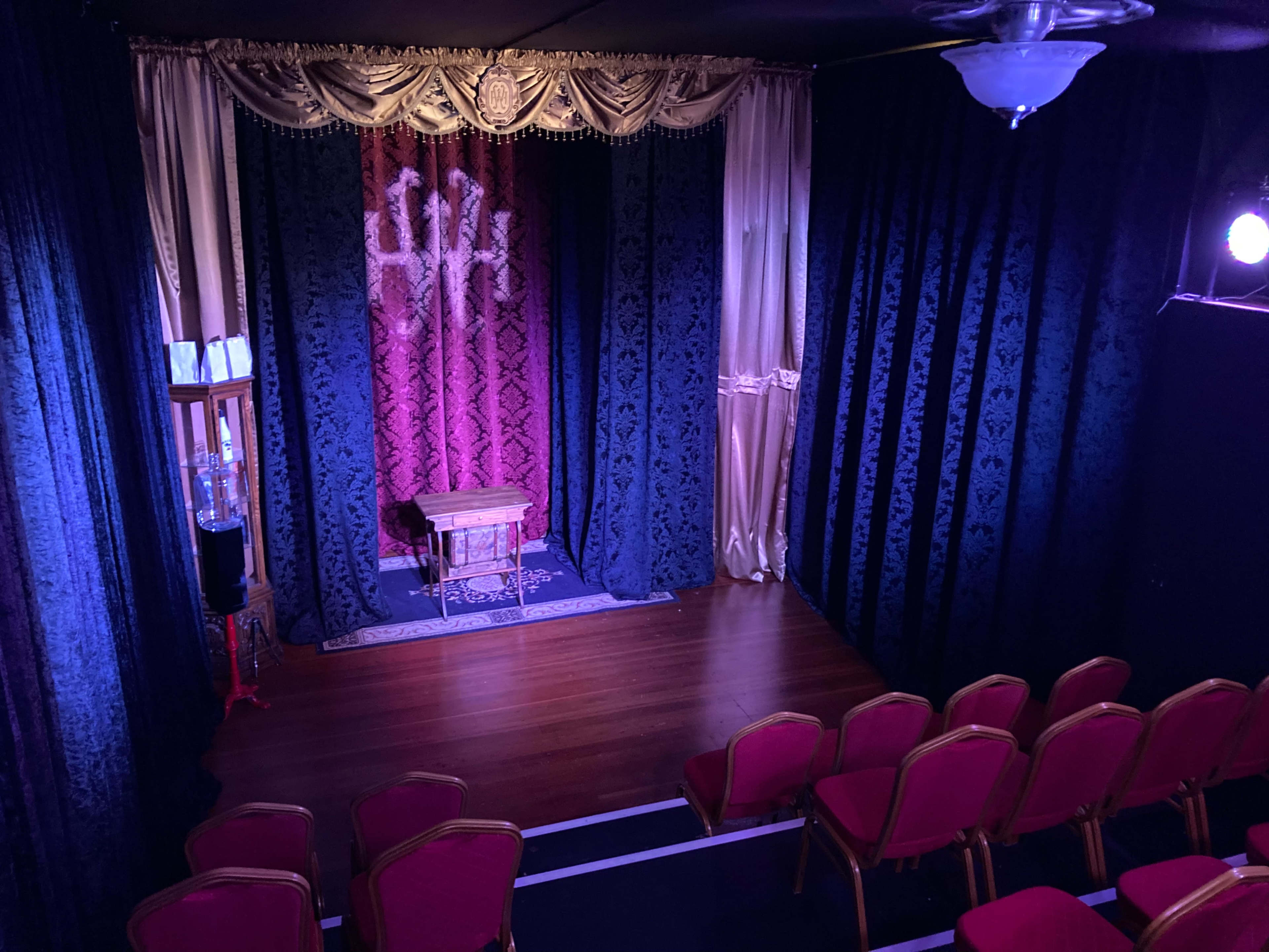 A small theater stage with red and black curtains, a wooden table in the center, and rows of red chairs facing the stage.