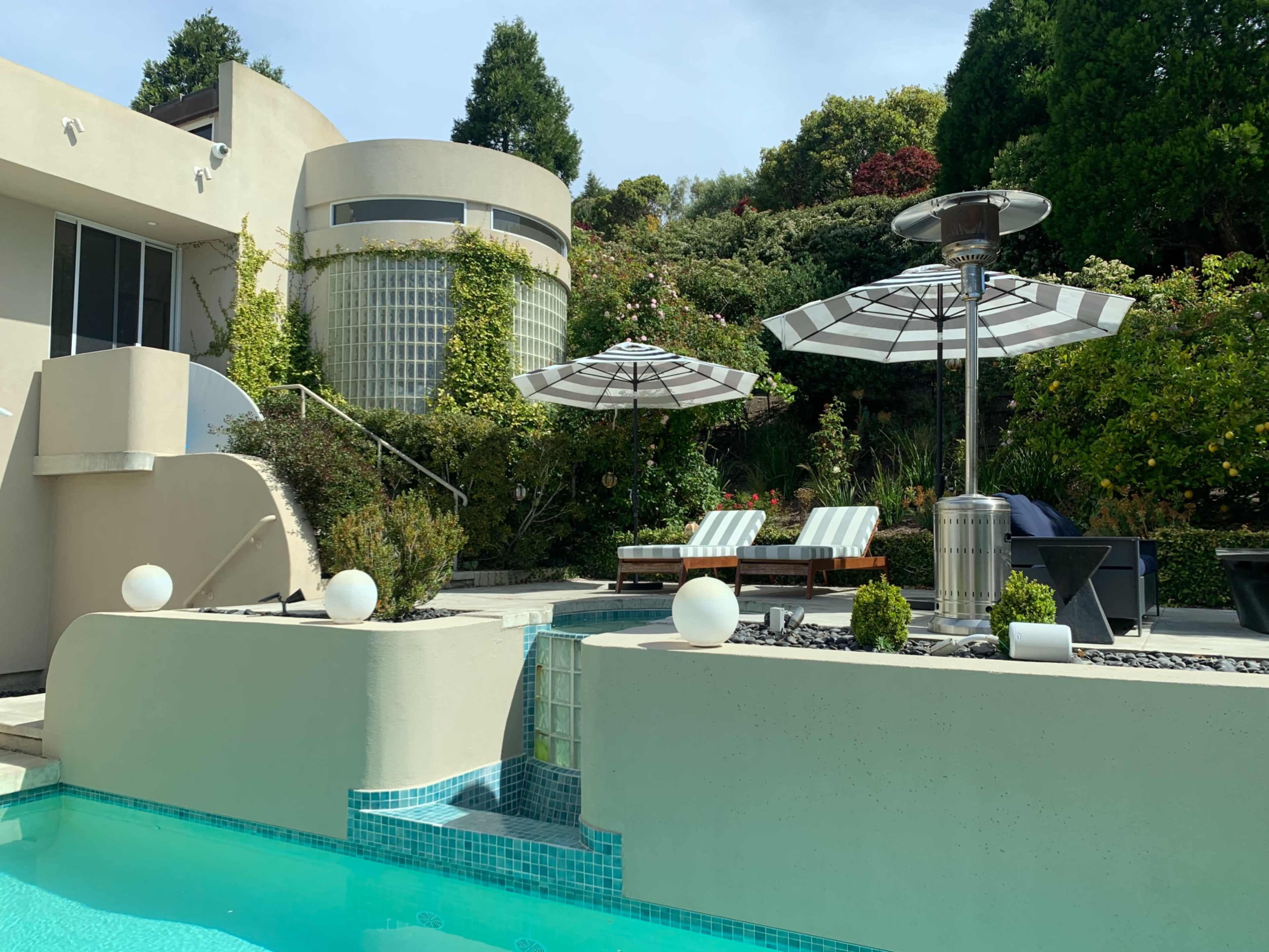 A modern pool area features striped umbrellas above lounge chairs, surrounded by landscaped greenery and water features.