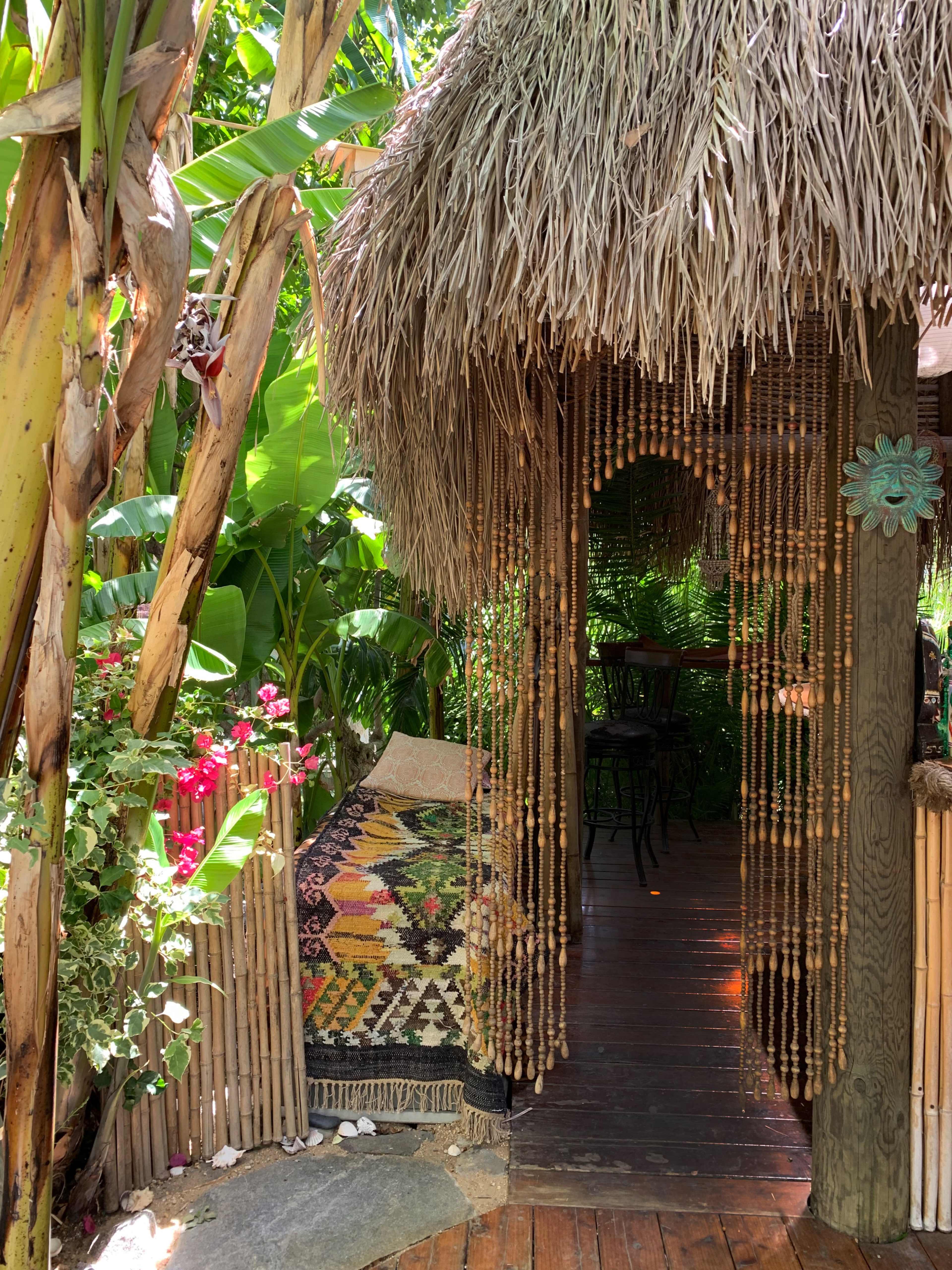 A thatched roof entrance draped with beaded curtains leads into a lush, green garden area adorned with vibrant flowers and a patterned rug.