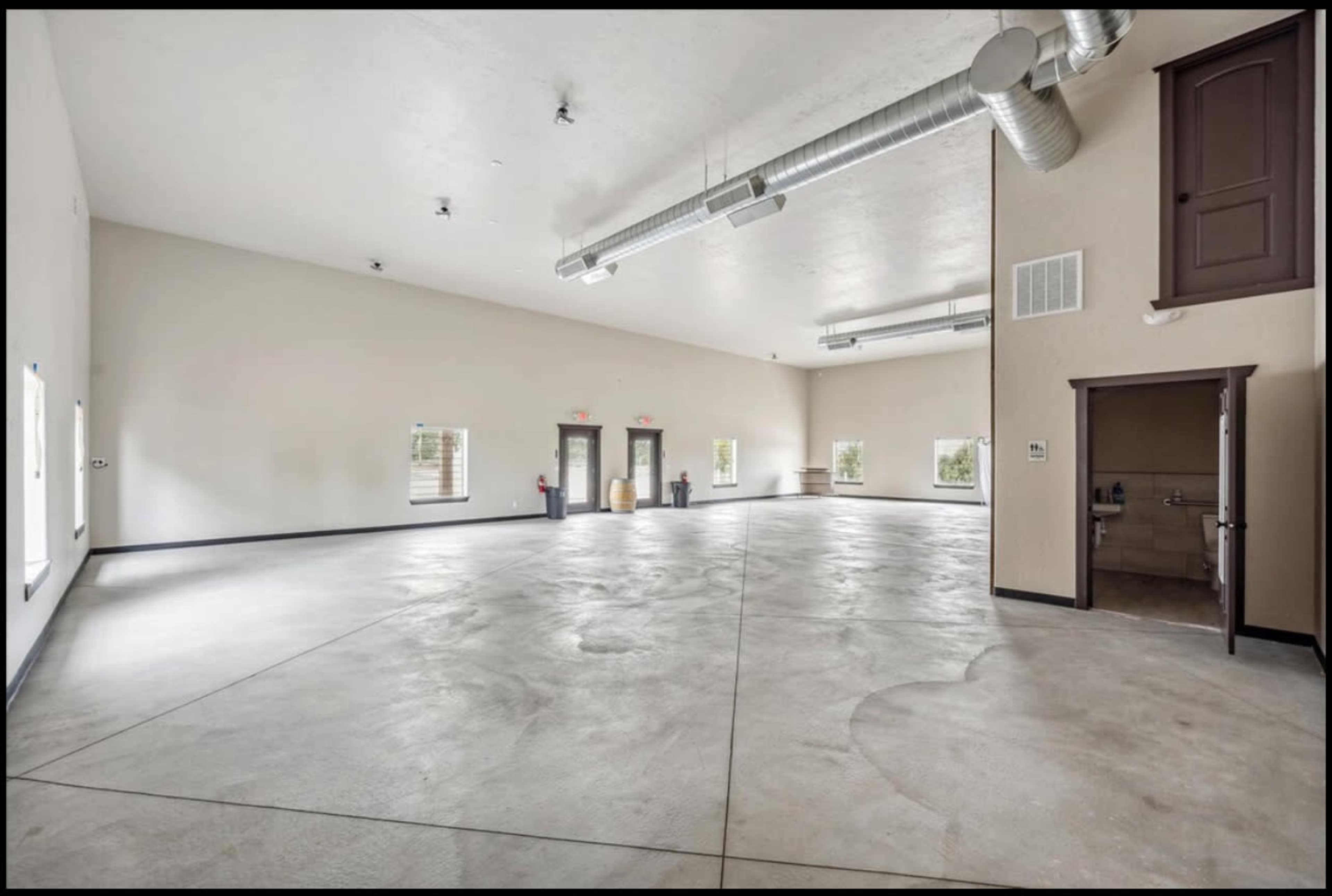The image shows a large, empty room with gray concrete floors, plain white walls, and several windows along the sides.