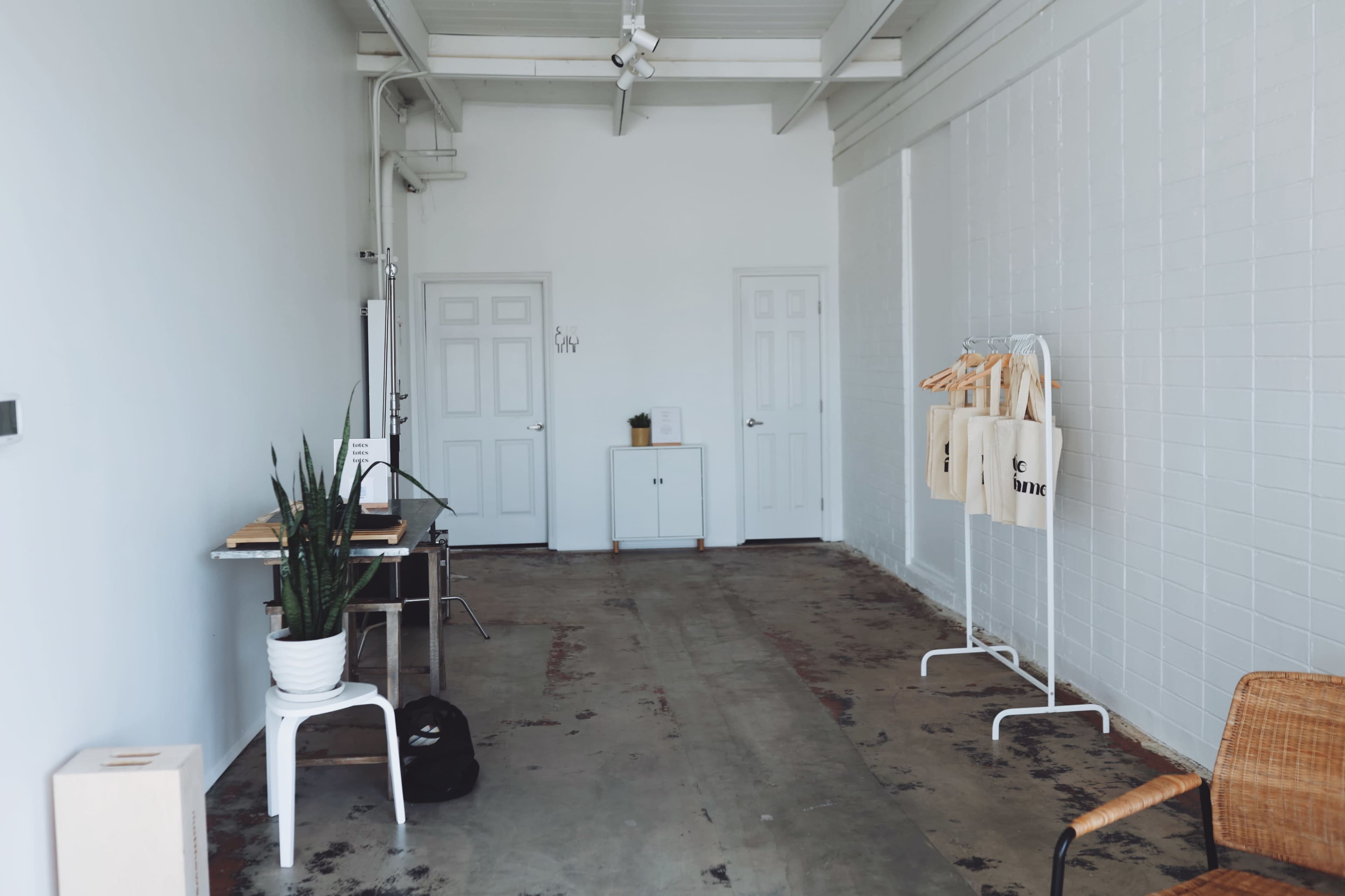 A minimalist interior space with white walls, two doors, a table, a display rack with tote bags, a potted plant, and a chair.