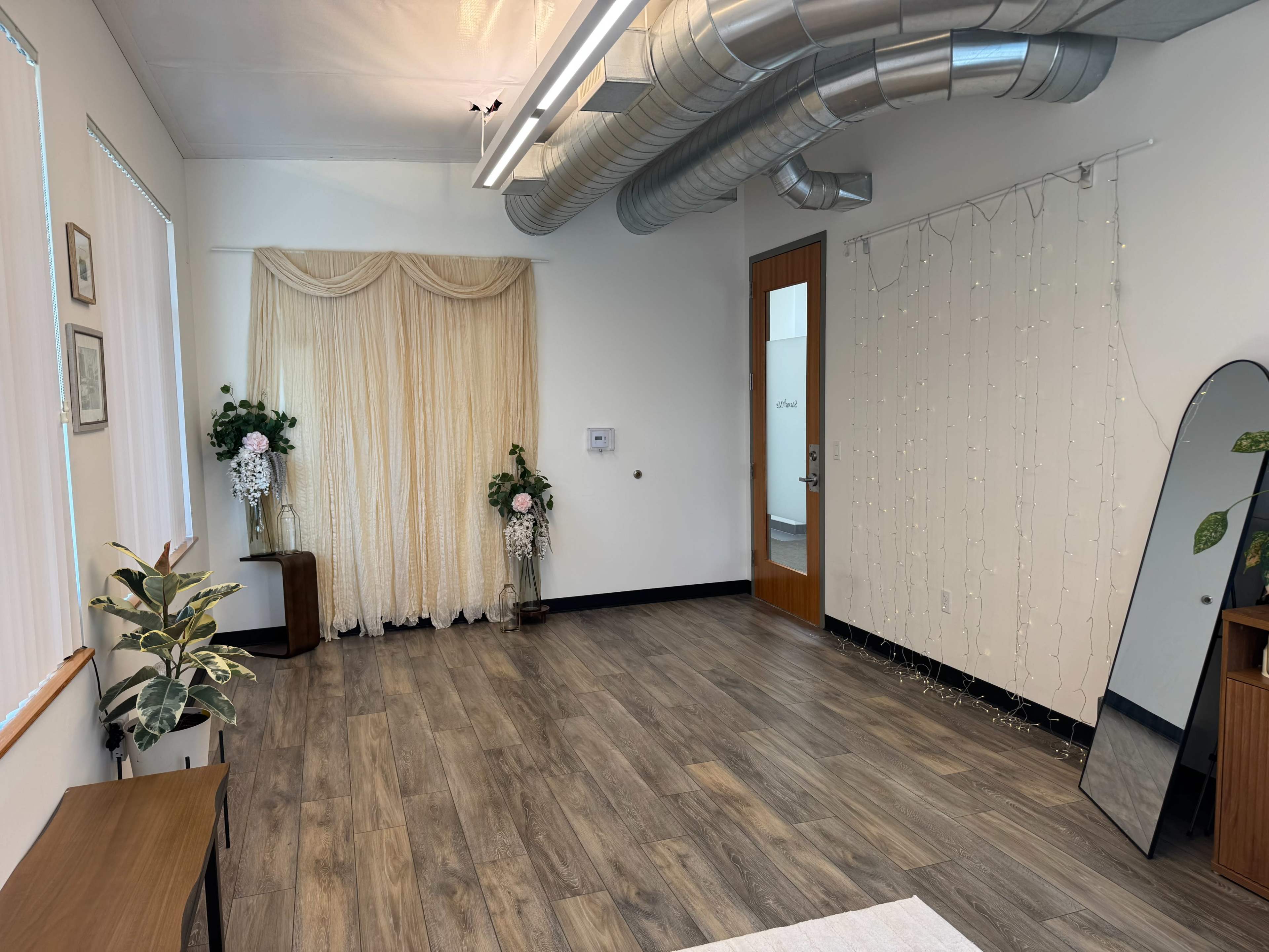 The image shows a spacious, well-lit room with wooden flooring, decorated with a pair of floral arrangements, a large mirror, and draped curtains.