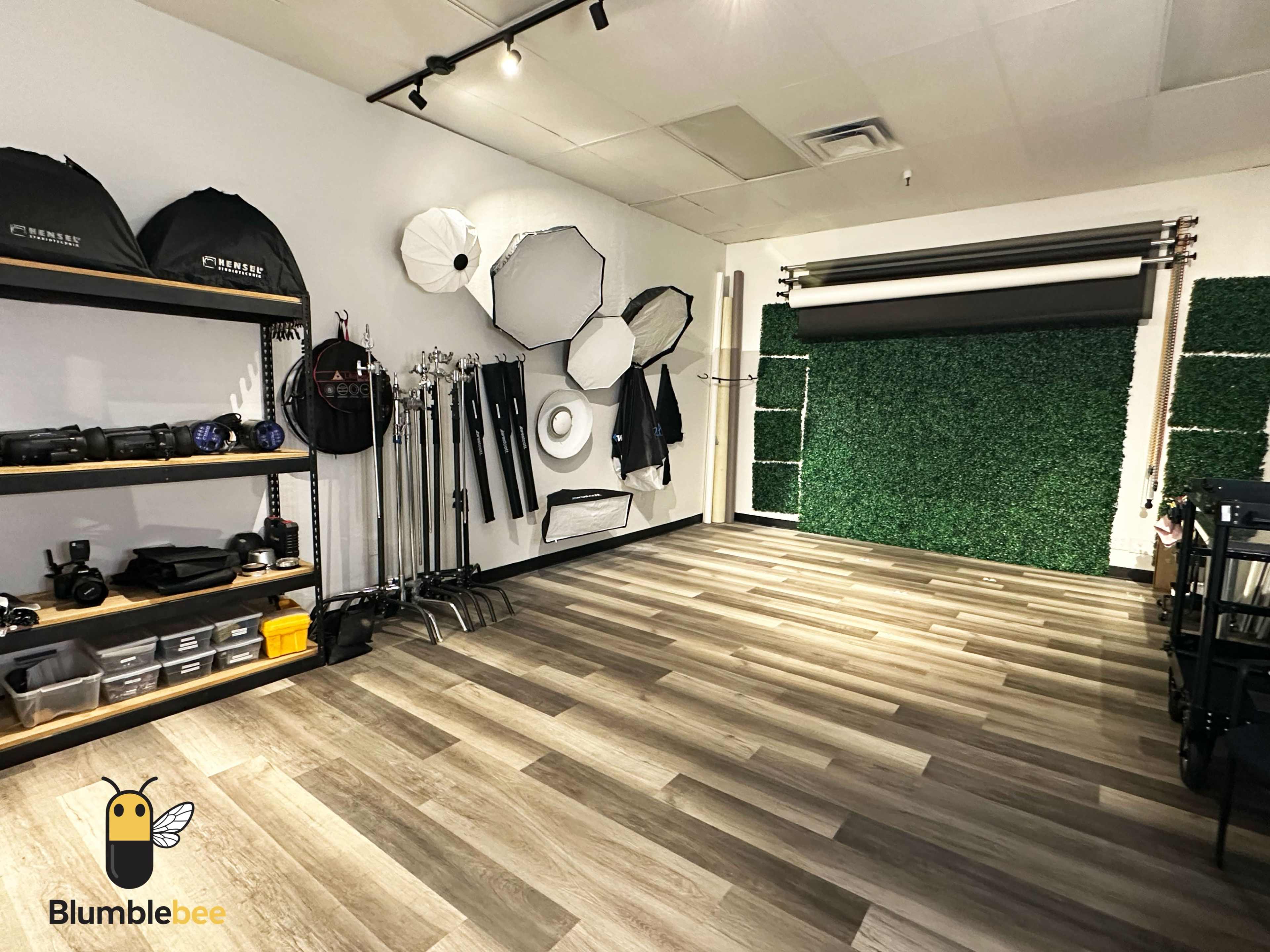 The image shows a photography studio featuring equipment shelves on the left, seamless backdrop rolls on the right, and a green textured wall.