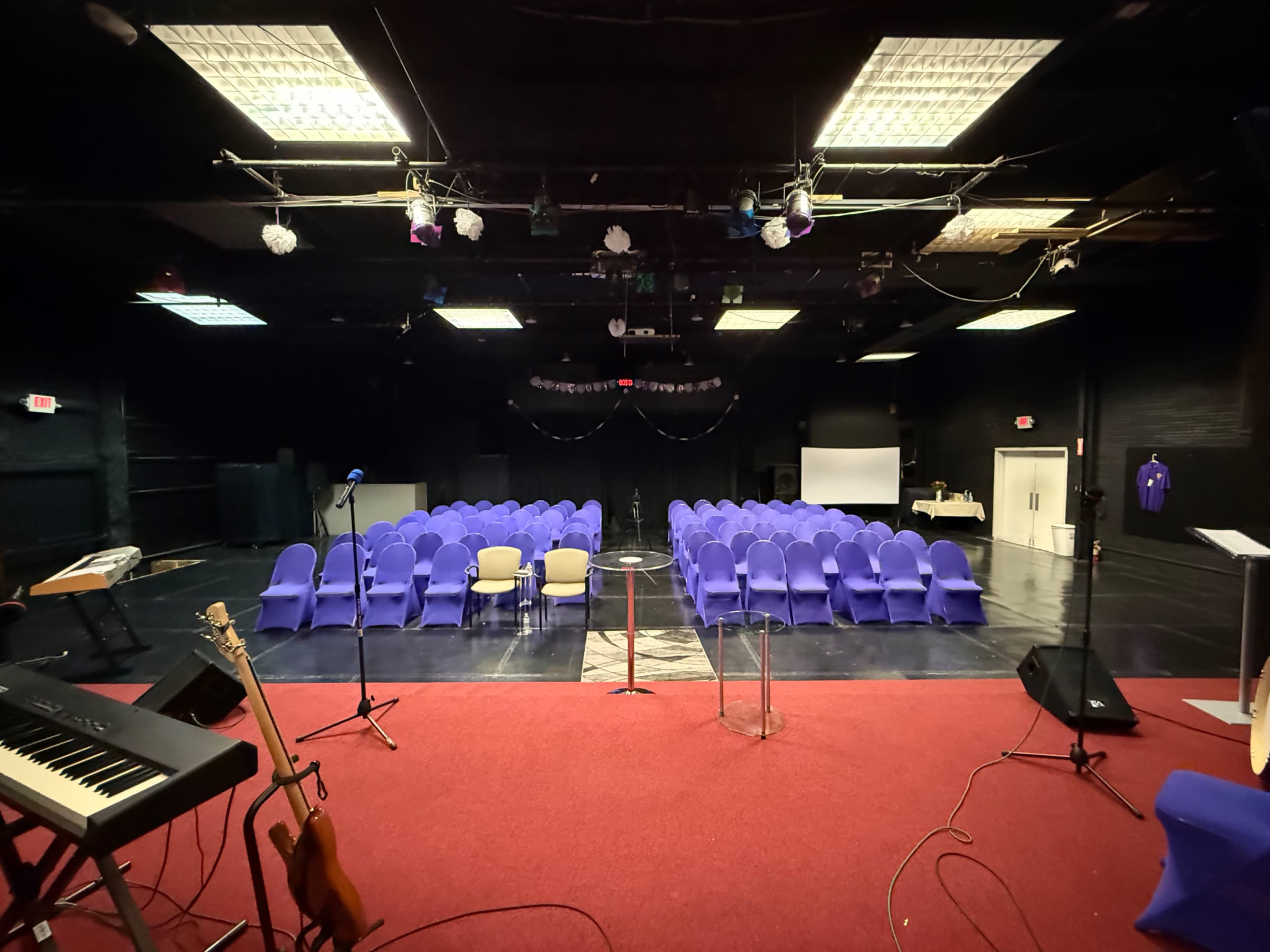 The image shows a setup for an event in a dimly lit auditorium with rows of purple chairs arranged in front of a stage.