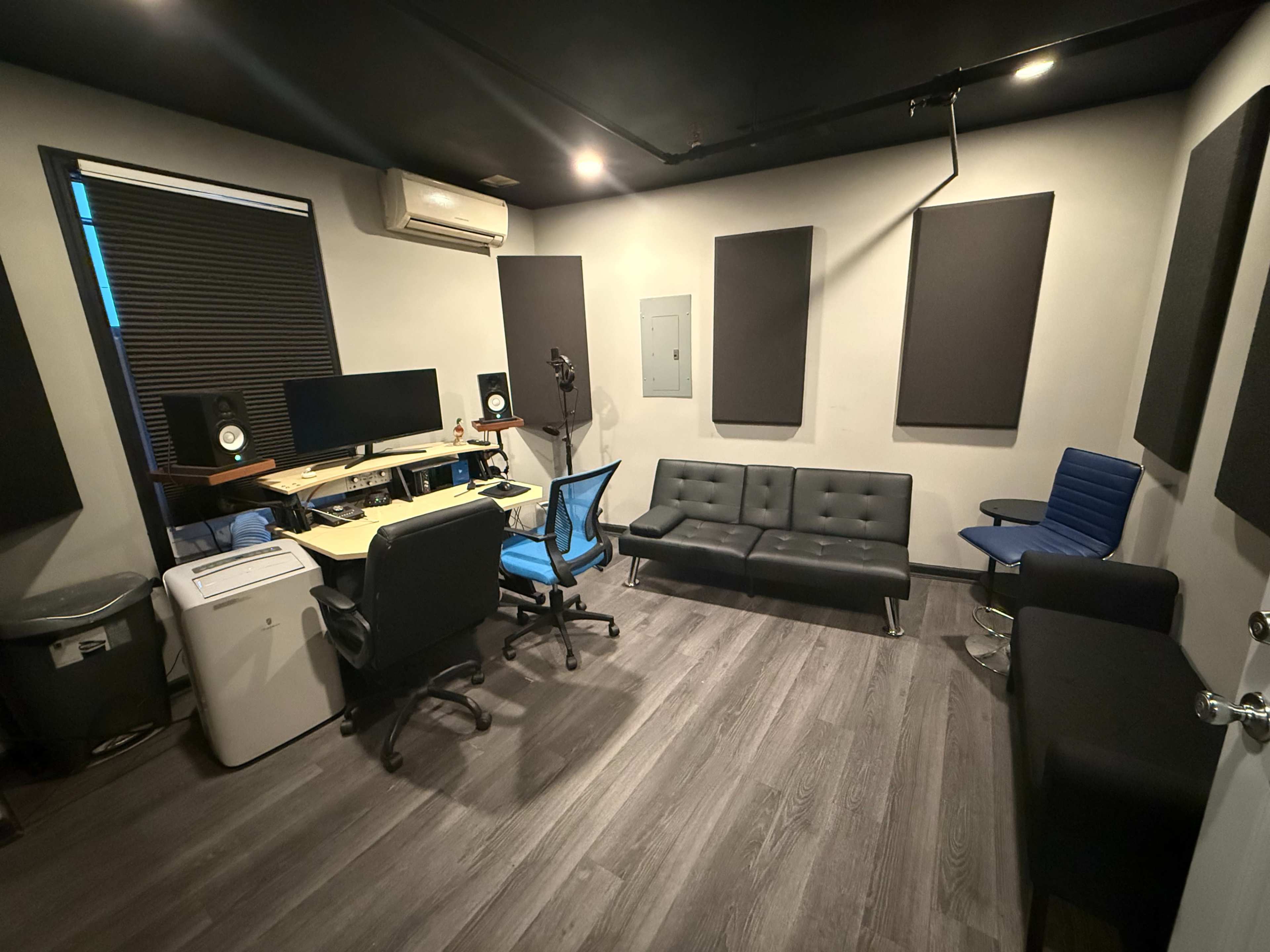 The image shows a small recording studio with soundproofing panels, a desk with a computer and speakers, and seating areas.