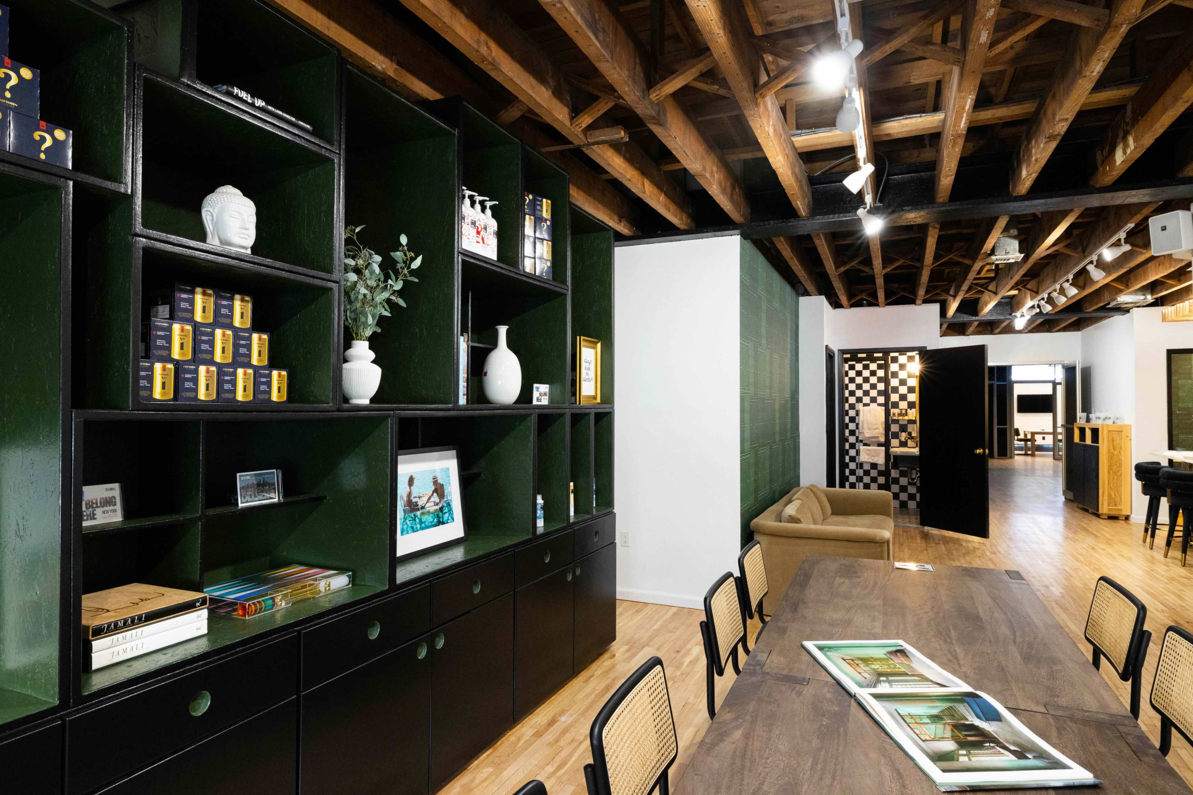 The image shows an interior space with a dark green shelving unit filled with decor and books, a long wooden table, and a view of a room with checkered flooring in the background.