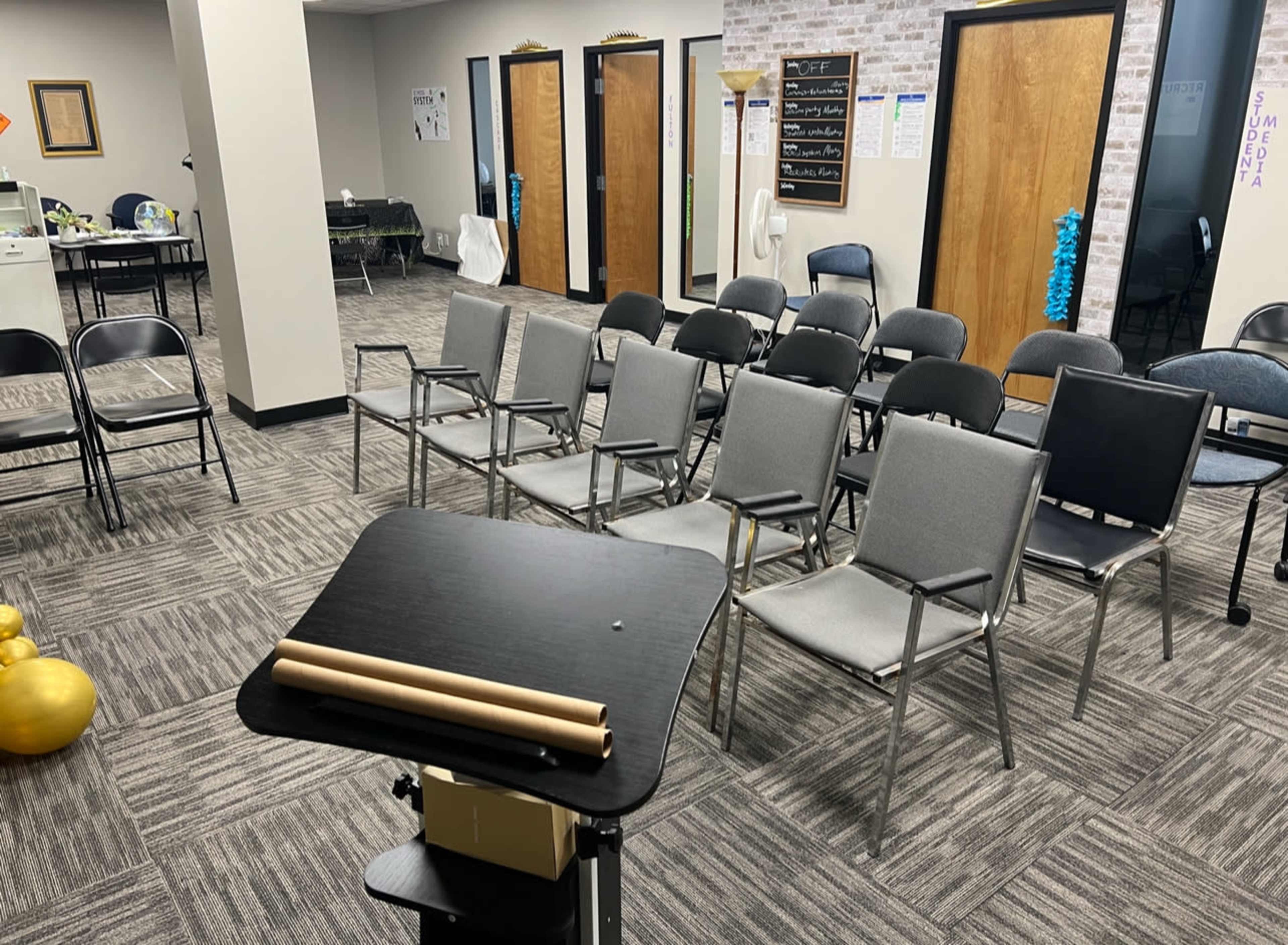 A room with several rows of gray chairs arranged in front of a black podium, surrounded by light-colored walls and a patterned carpet.