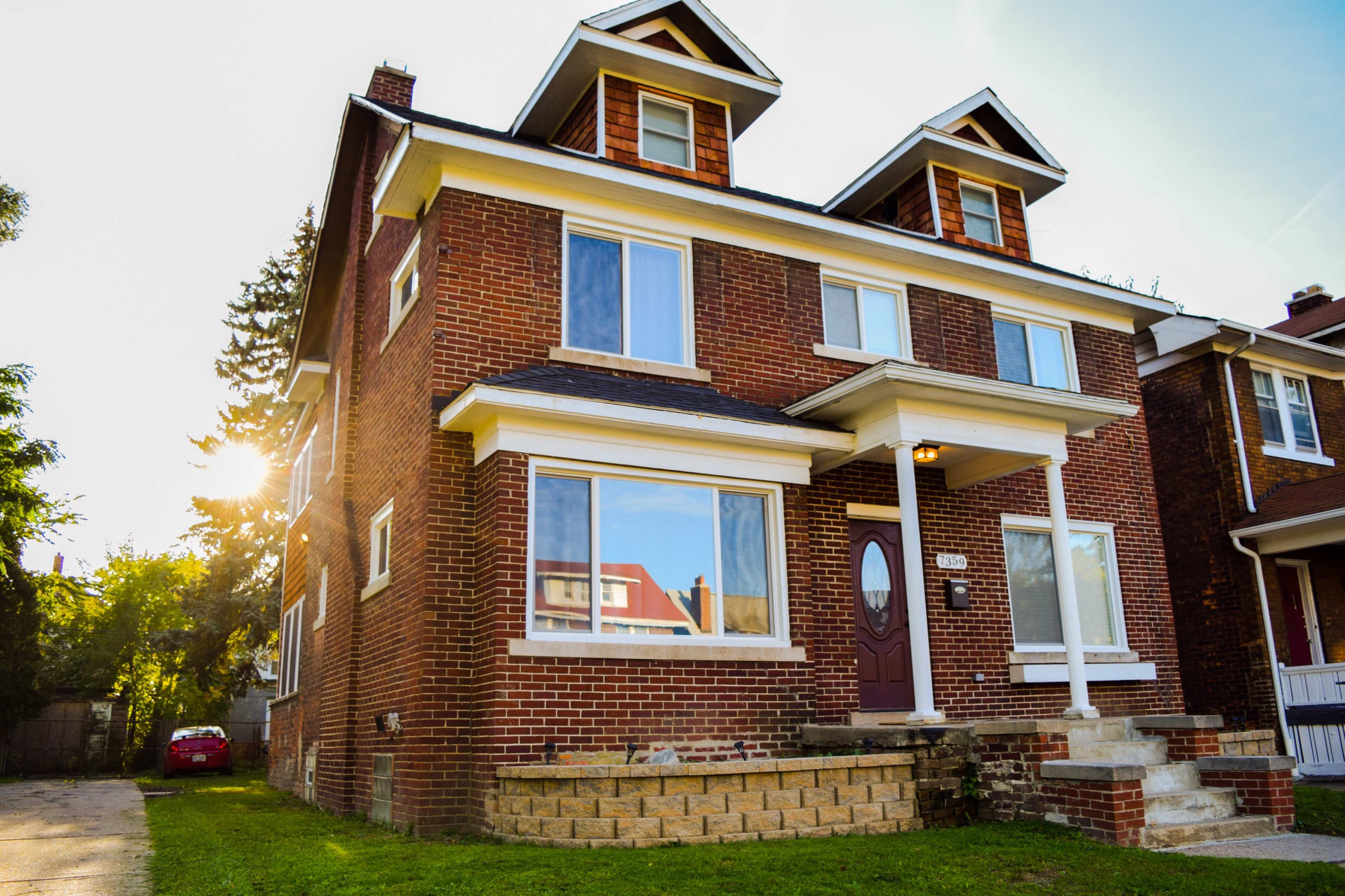A red brick house with two dormer windows and a large front porch is situated on a grassy lawn.