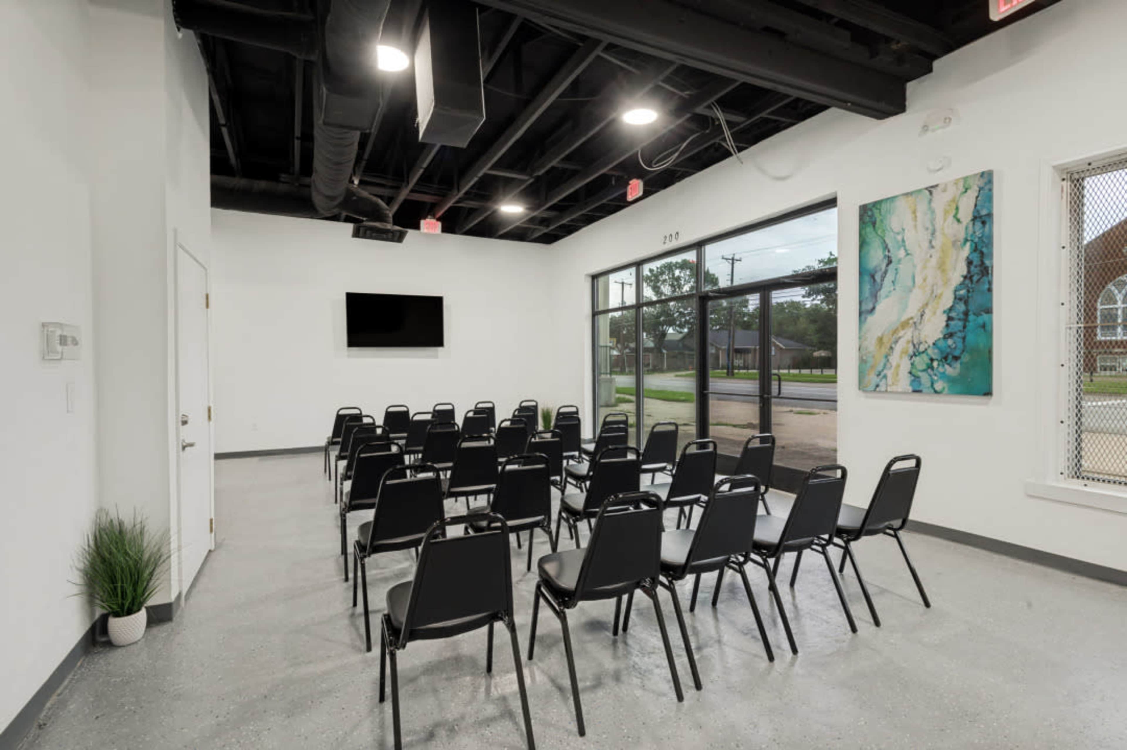 The image shows a modern, sparsely furnished room set up with rows of black chairs facing a wall-mounted television and a large window overlooking a street.