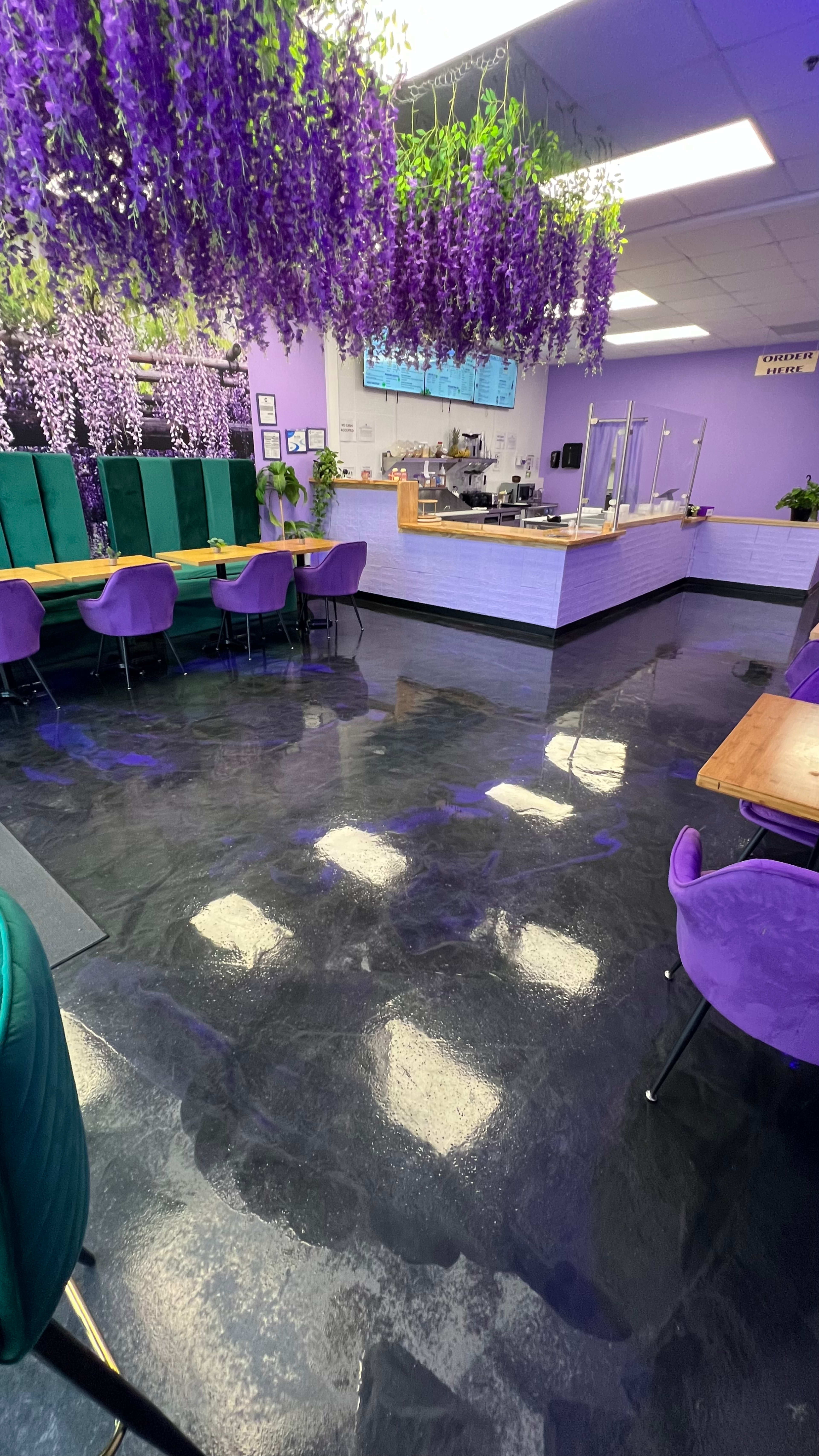 The image shows an interior of a café decorated with purple wisteria flowers hanging from the ceiling, purple chairs, and a counter area in the background.