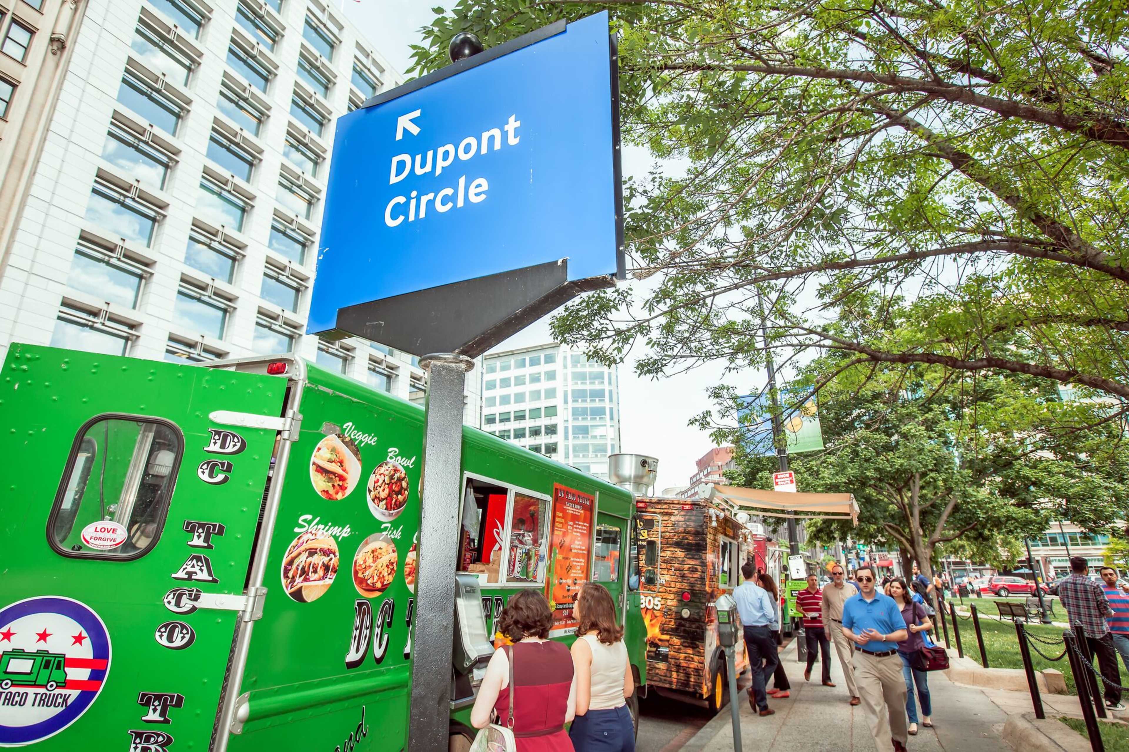 A green food truck and a brown food truck are stationed near a sign pointing to Dupont Circle, with several people walking by on a busy sidewalk.