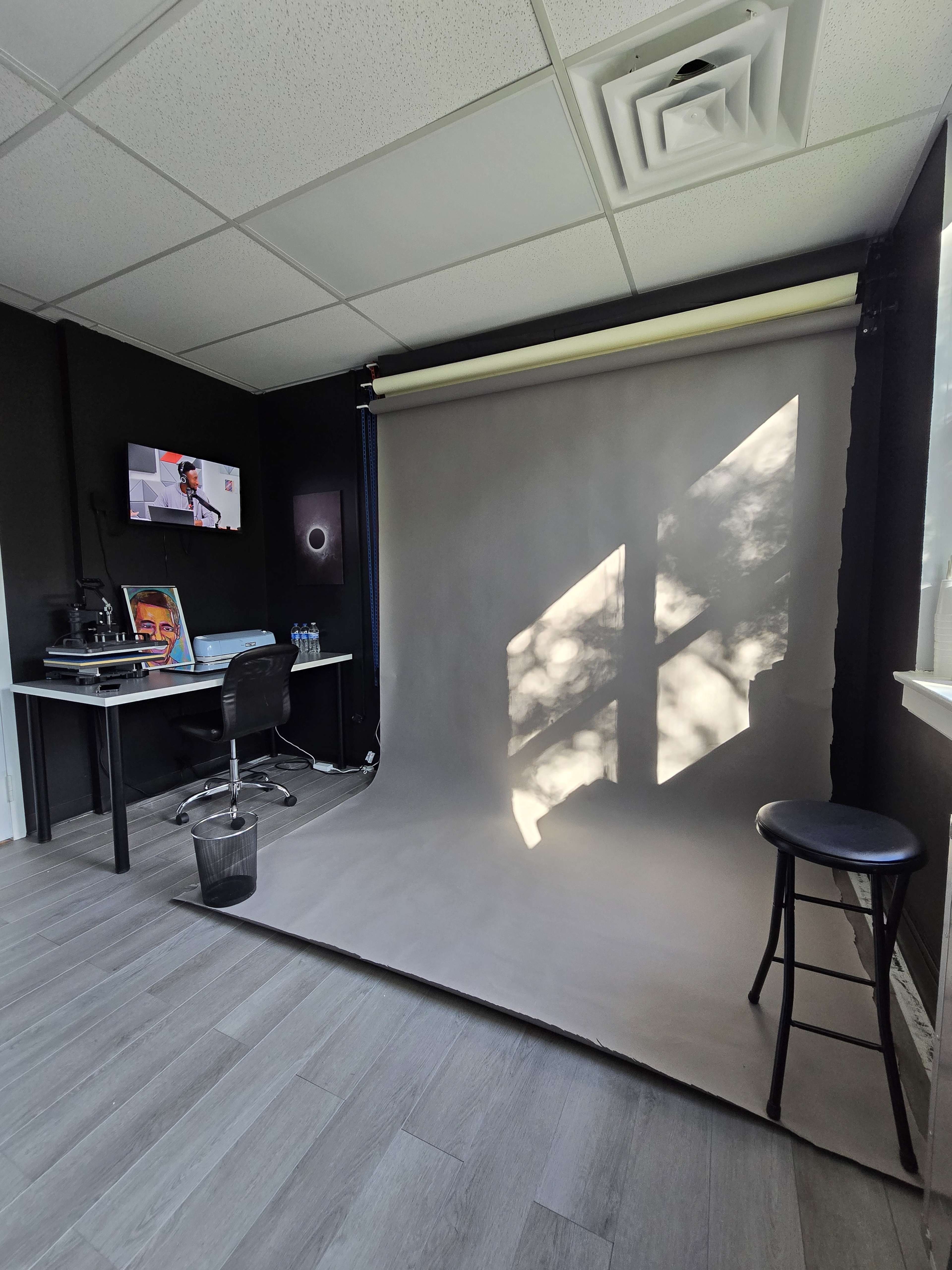 A photography studio with a gray backdrop, a chair, a desk with a computer and monitor, and natural light casting shadows on the floor.