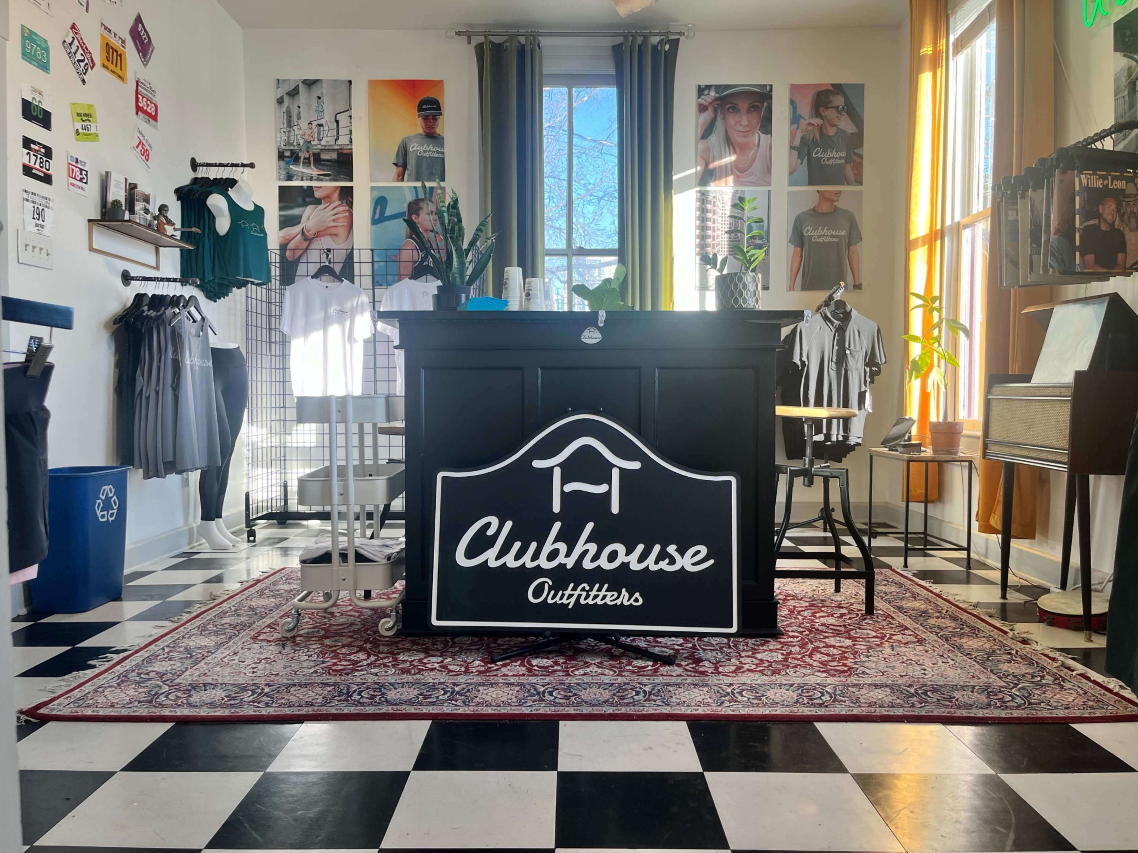 An interior retail space with a checkered floor, a counter featuring a neon sign that reads "Clubhouse Outfitters," and various clothing items displayed on racks and walls.