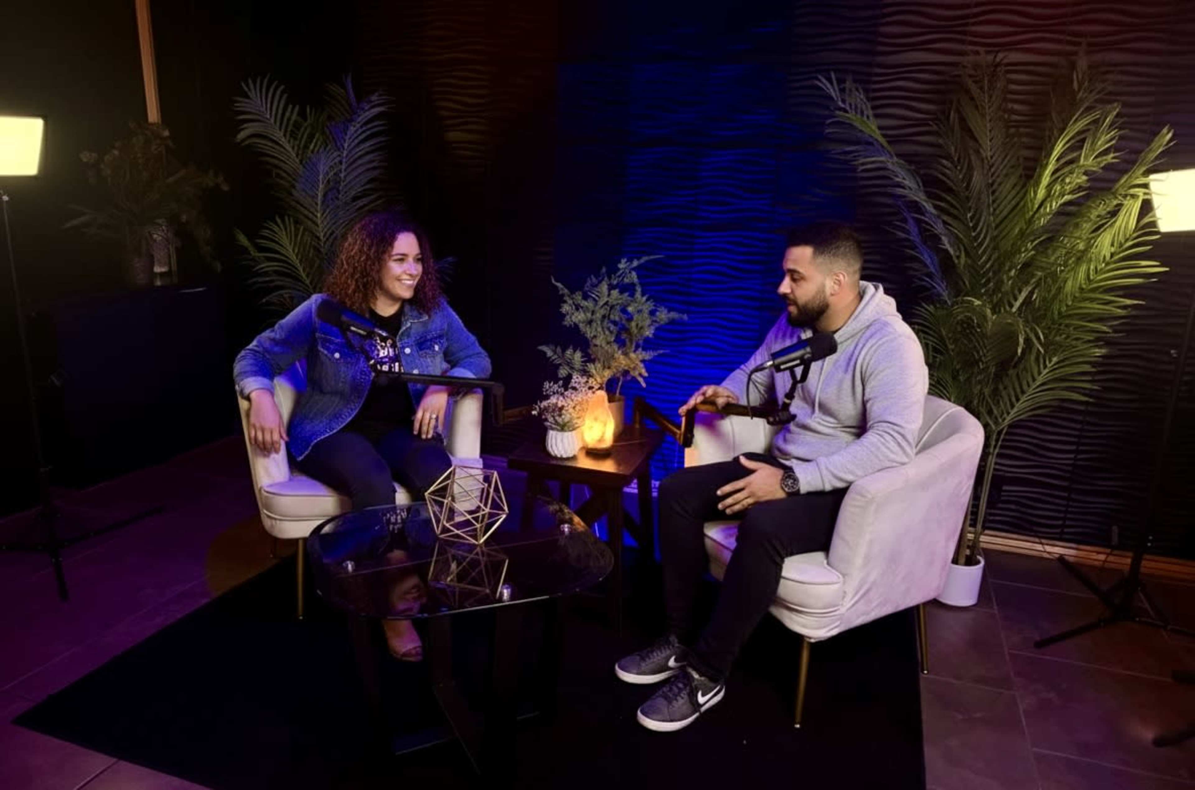 A man and a woman are sitting in a modern interview setting with a dark backdrop and plants, engaged in conversation.