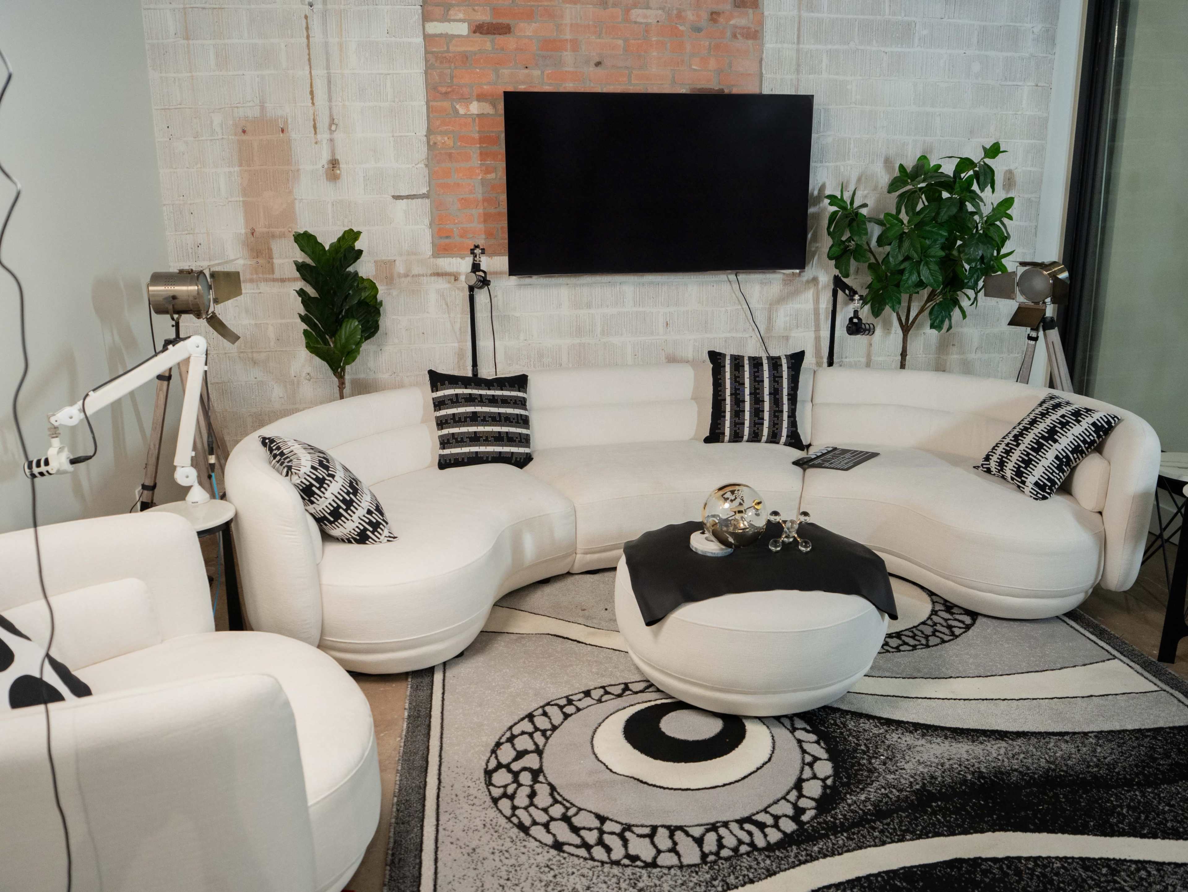 The image shows a modern living room with a curved white sofa, black and white patterned pillows, a round coffee table on a patterned rug, and a wall-mounted TV, complemented by potted plants and stylish floor lamps.